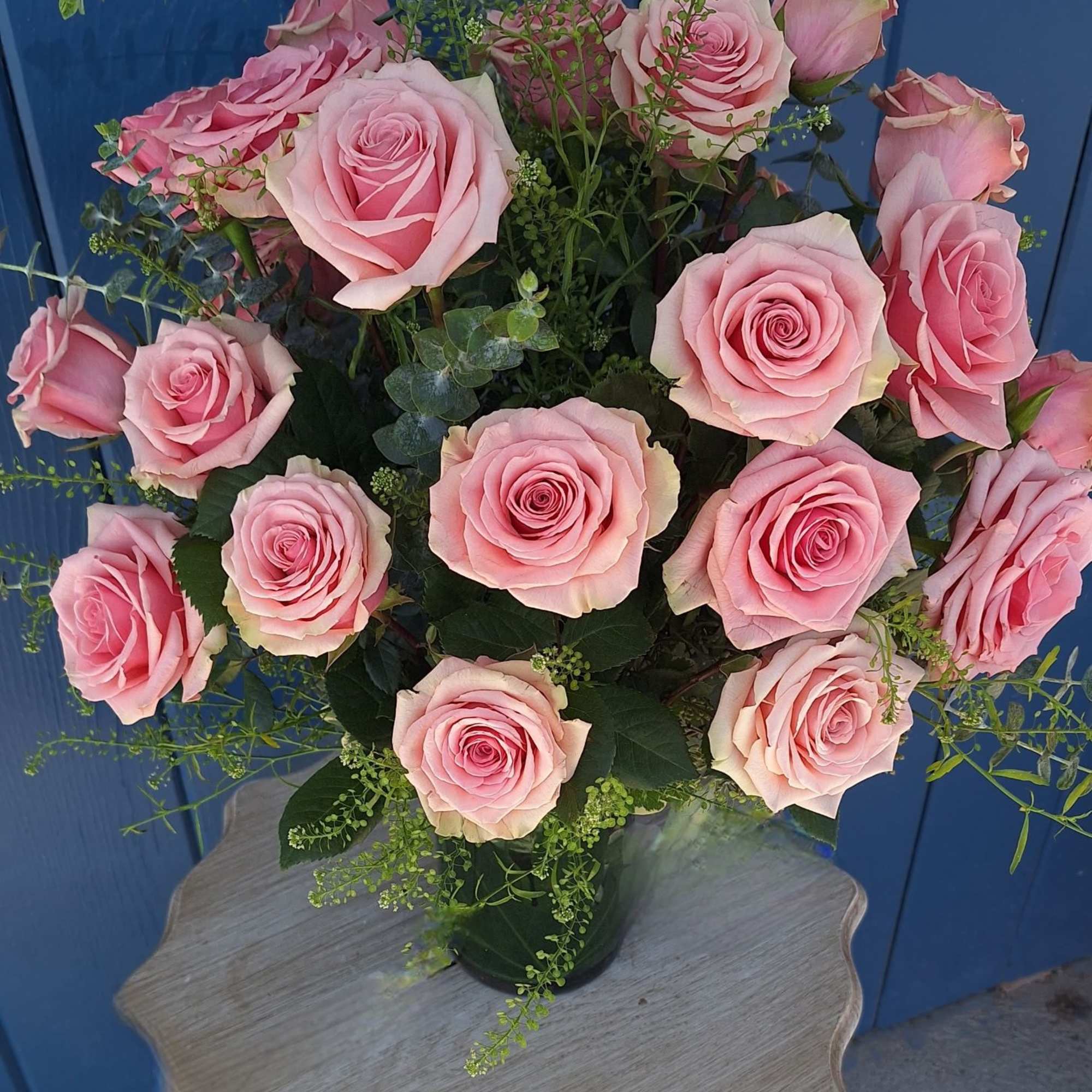 Arrangement of pink roses in a clear glass vase on a small wooden table against a blue wall