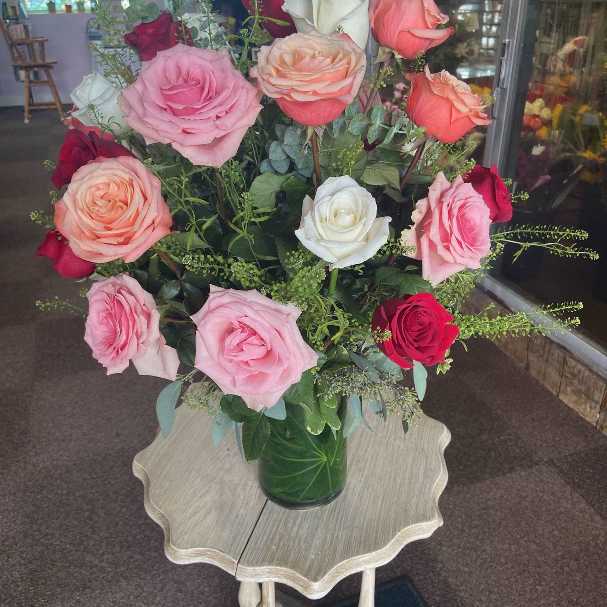 Tall arrangement of pink, peach, white, and red roses in a clear glass vase on a small wooden table.