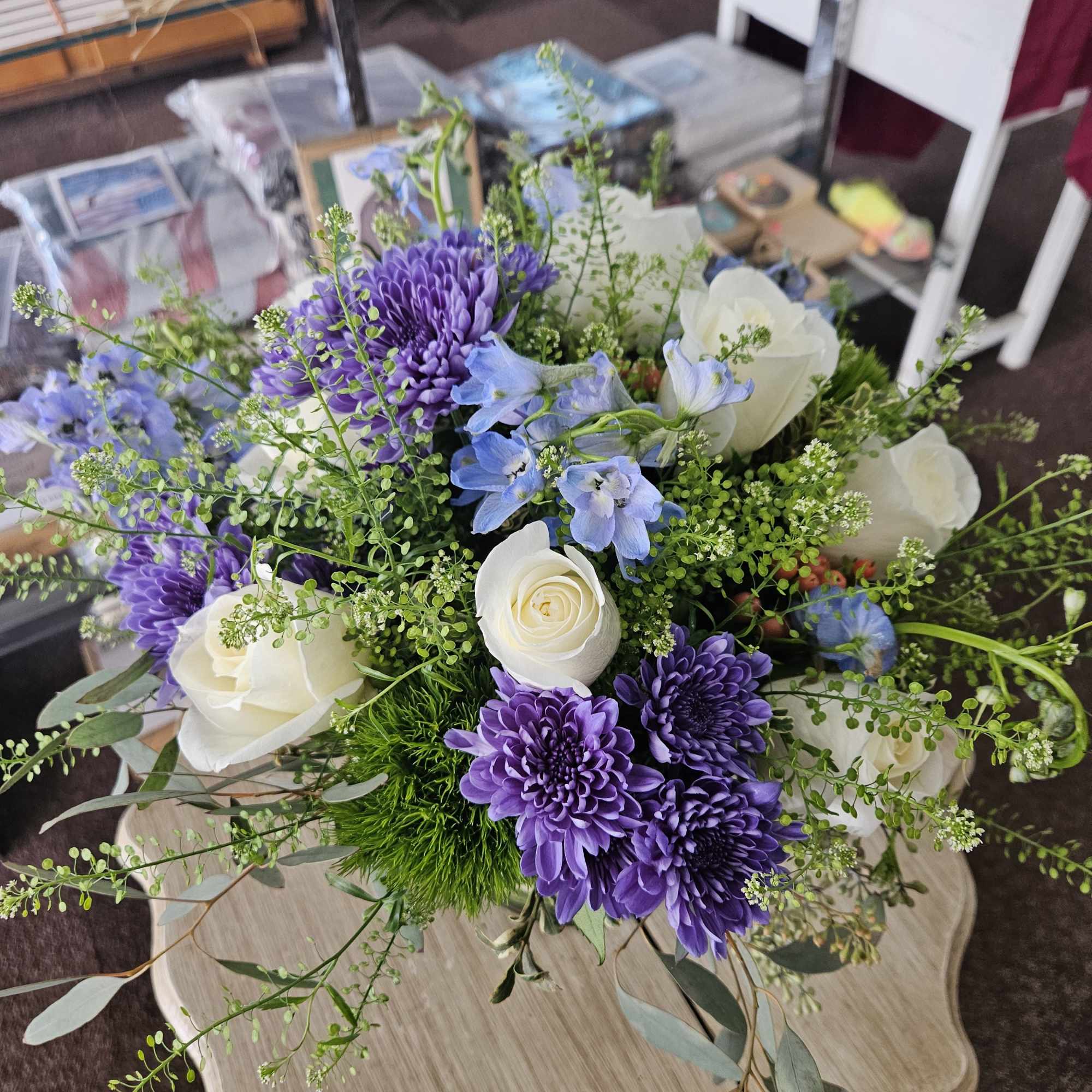 Arrangement of white roses, purple chrysanthemums, and blue flowers with airy greenery on a small wood table