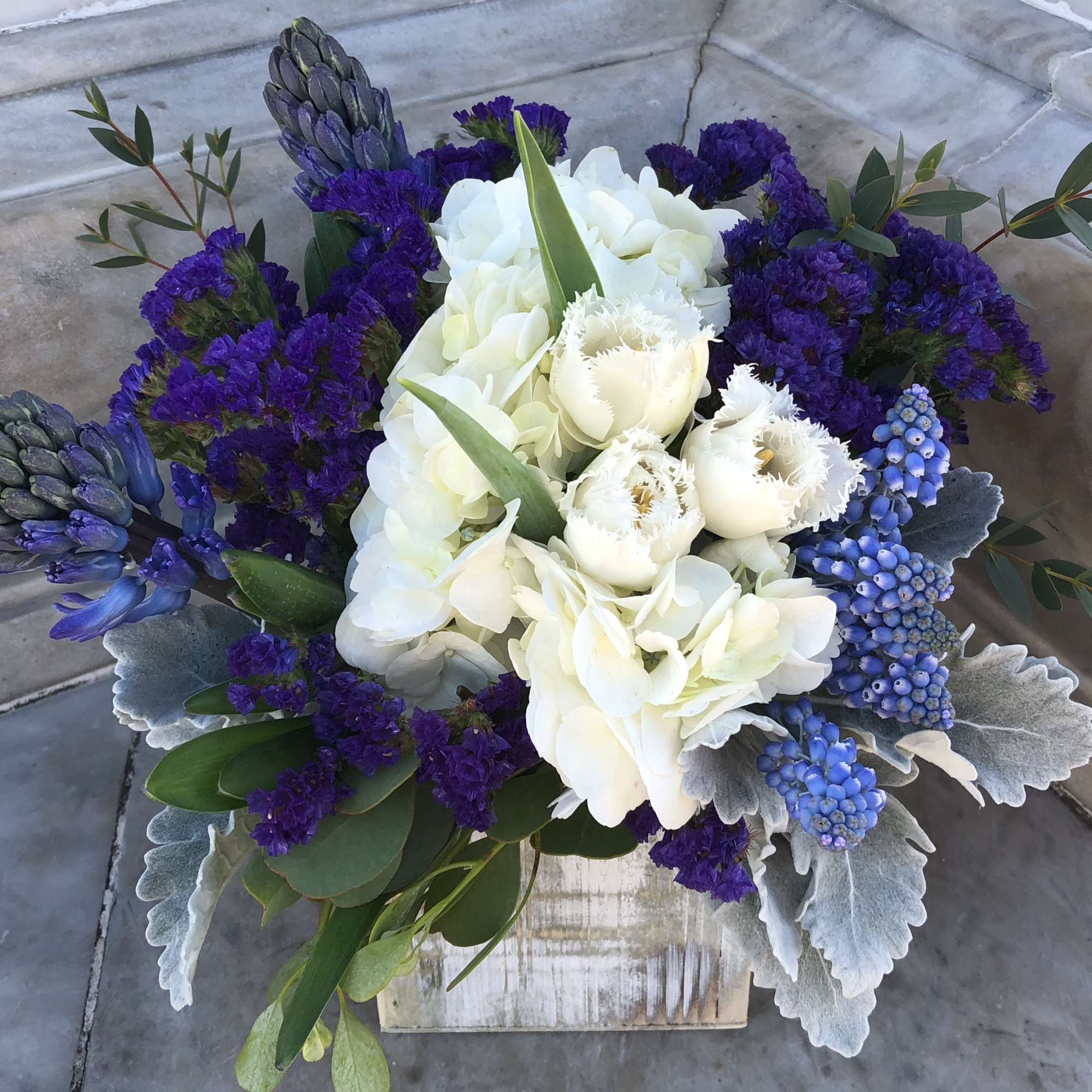 White and purple floral arrangement with blue grape hyacinths in a wooden container