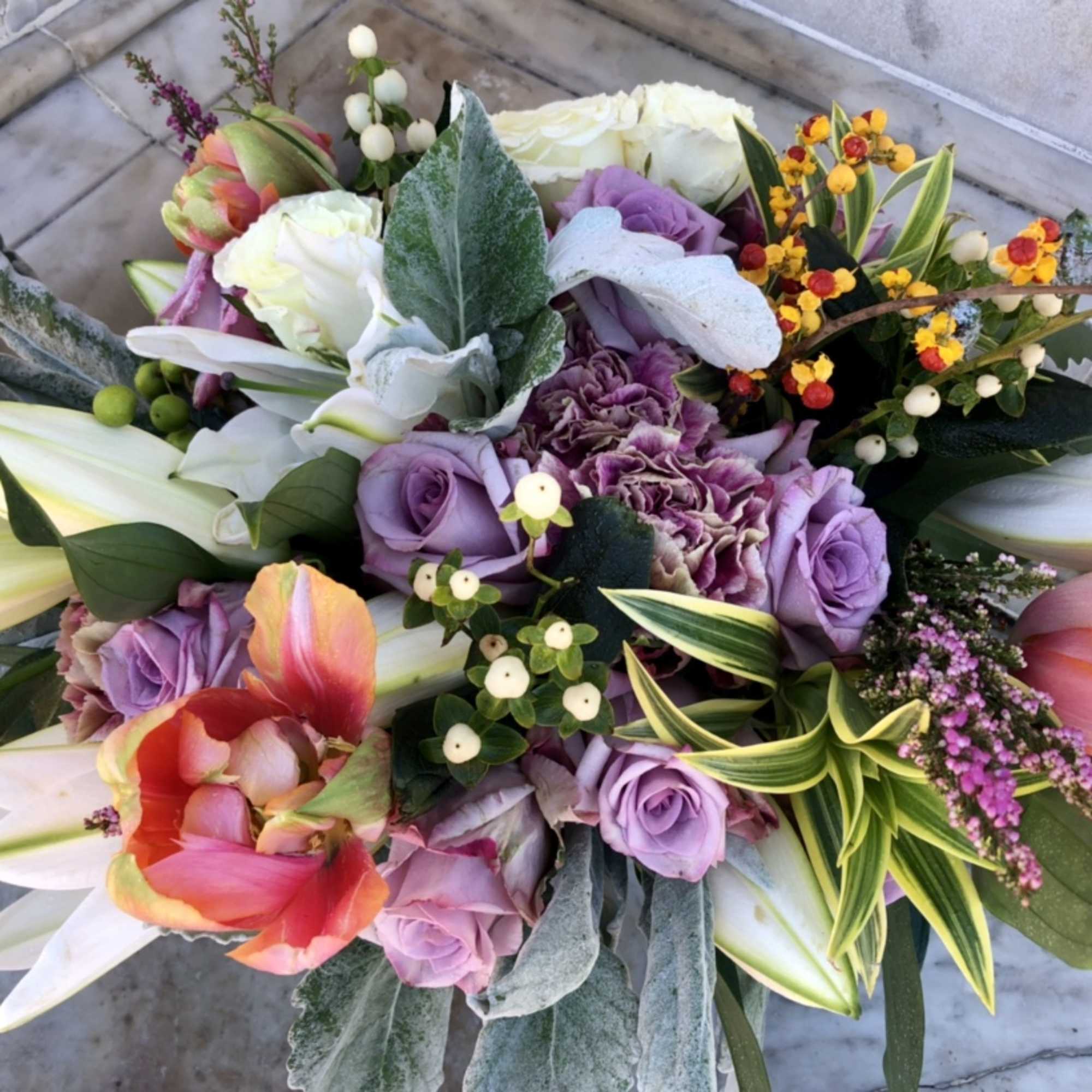 Colorful bouquet with white lilies, lavender roses, carnations, and tulips in warm pink and orange tones.