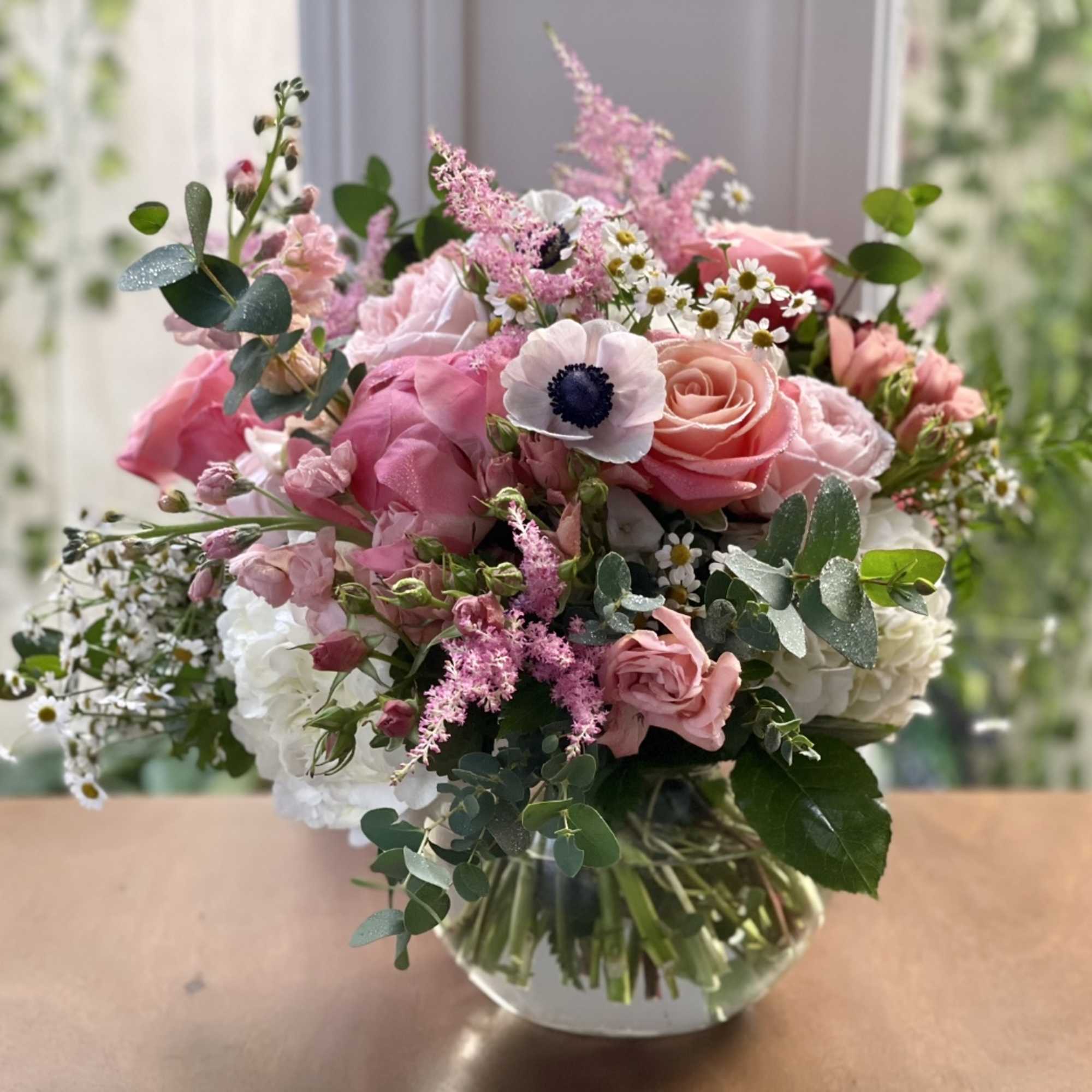 Pink and white mixed bouquet with roses and anemones in a round clear glass vase on a wooden surface.