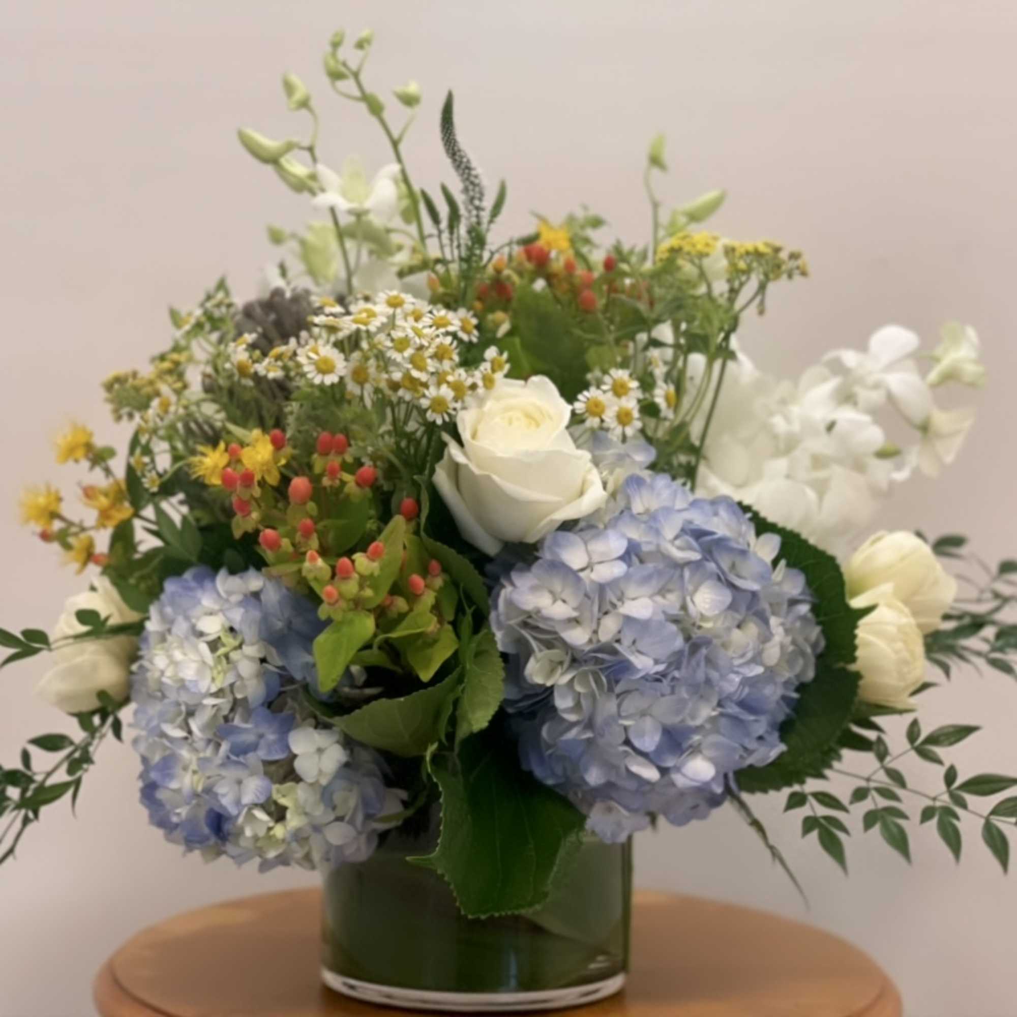Low glass vase of blue hydrangeas, white roses, and mixed yellow and white flowers