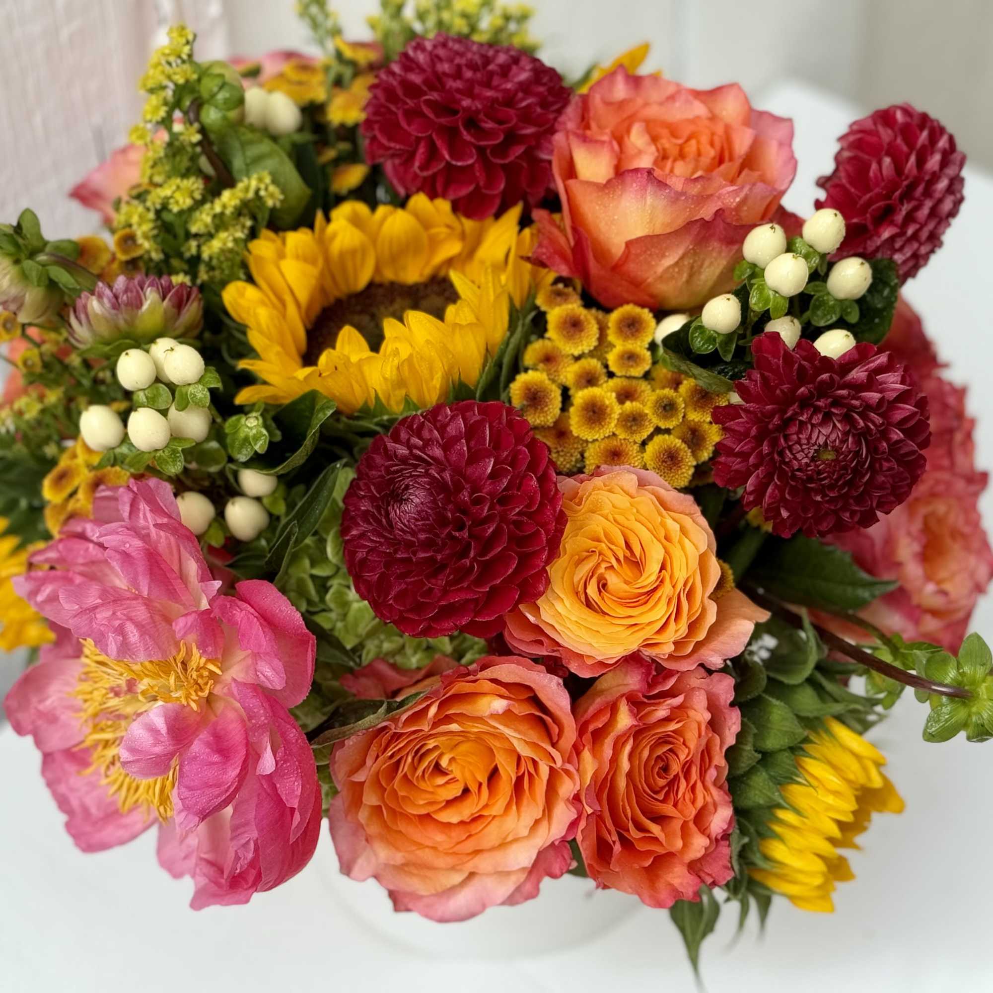 Low mixed arrangement of yellow sunflowers, orange roses, pink peonies, and burgundy dahlias in a white bowl vase