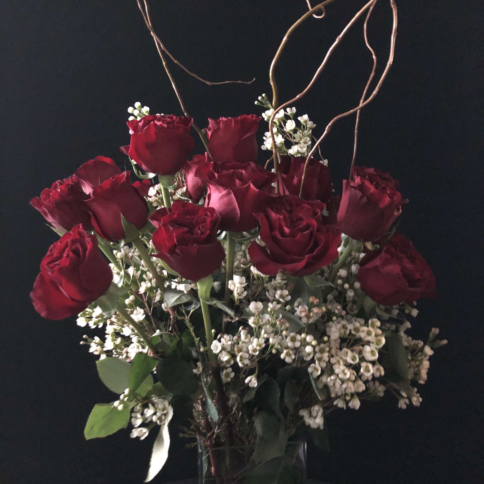 Red roses arranged in a clear vase with white filler flowers and curly branches