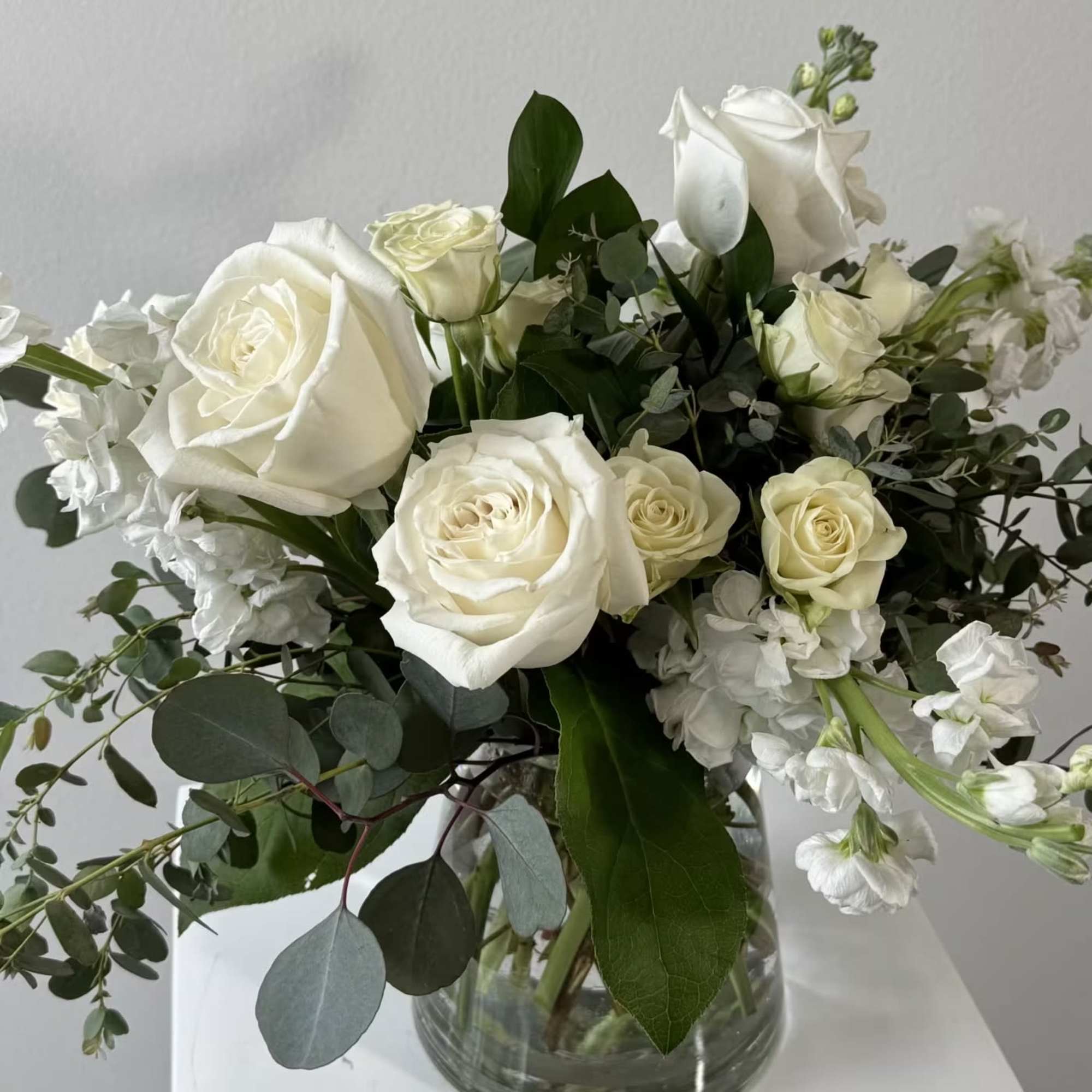 White roses and white stock in a clear glass vase with greenery.