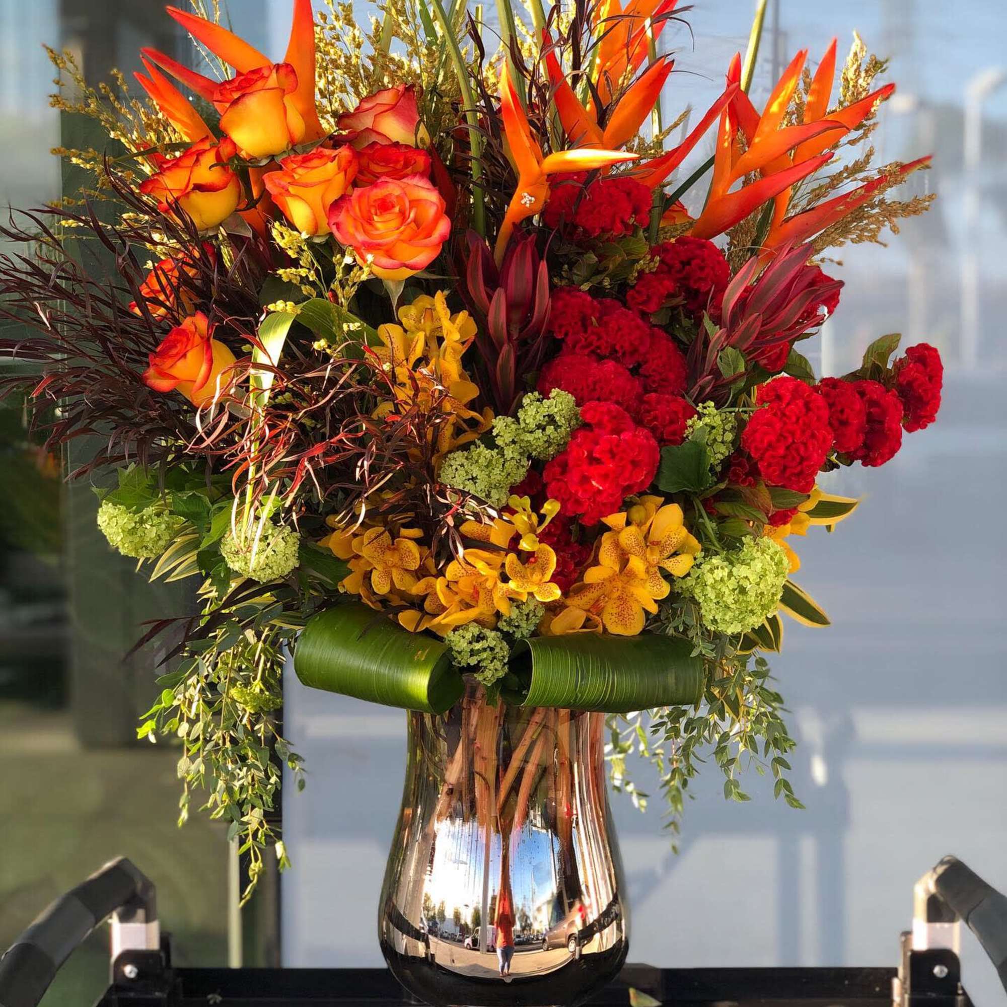 Tall bouquet of orange roses, red blooms, and orchids in a silver vase