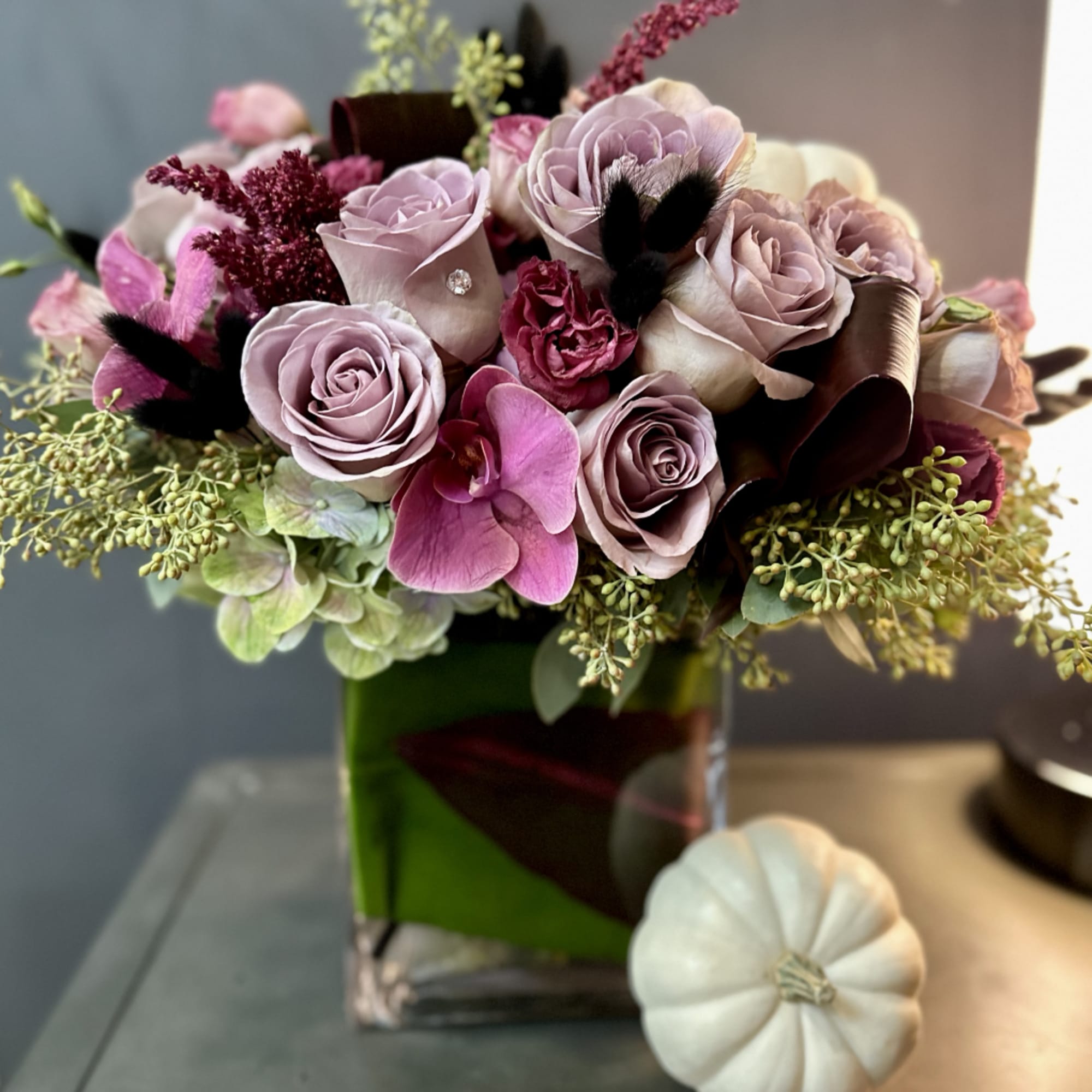 Pink and lavender rose bouquet in a green glass vase with a white pumpkin