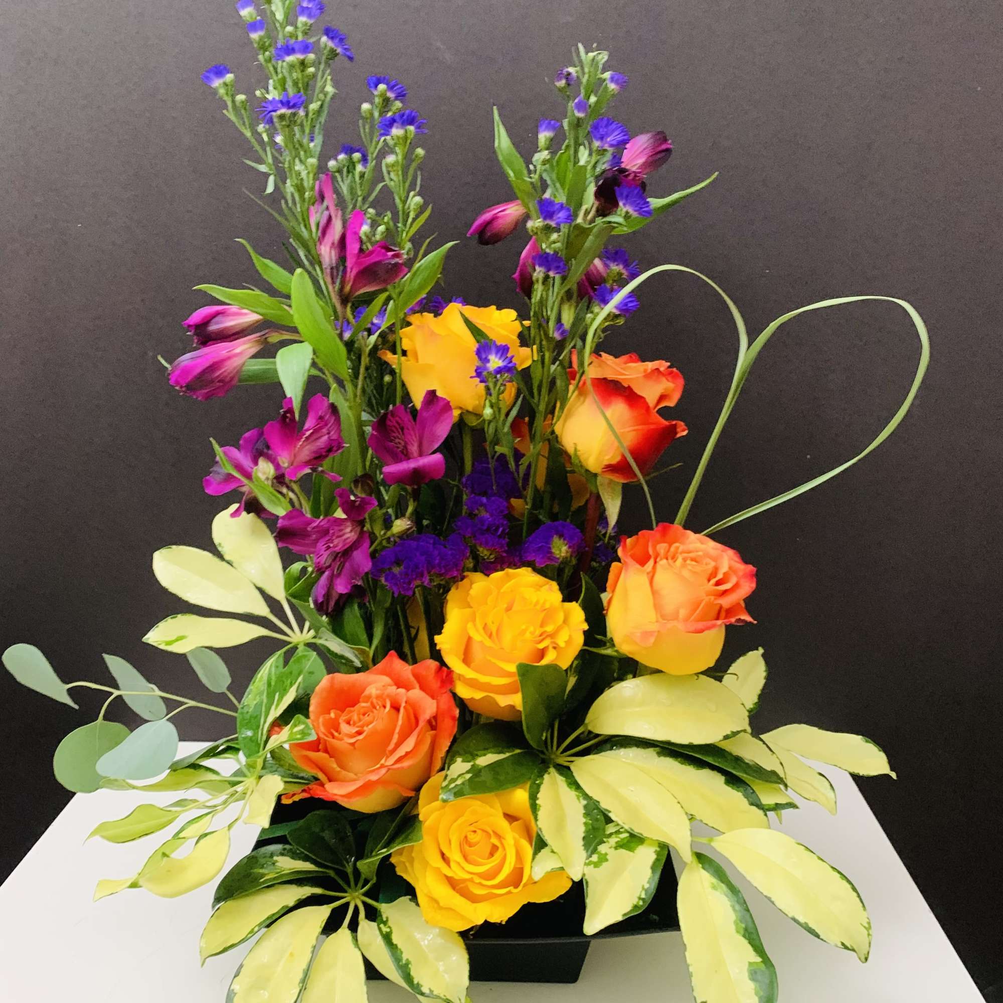 Mixed bouquet of orange and yellow roses with purple accent flowers in a black container
