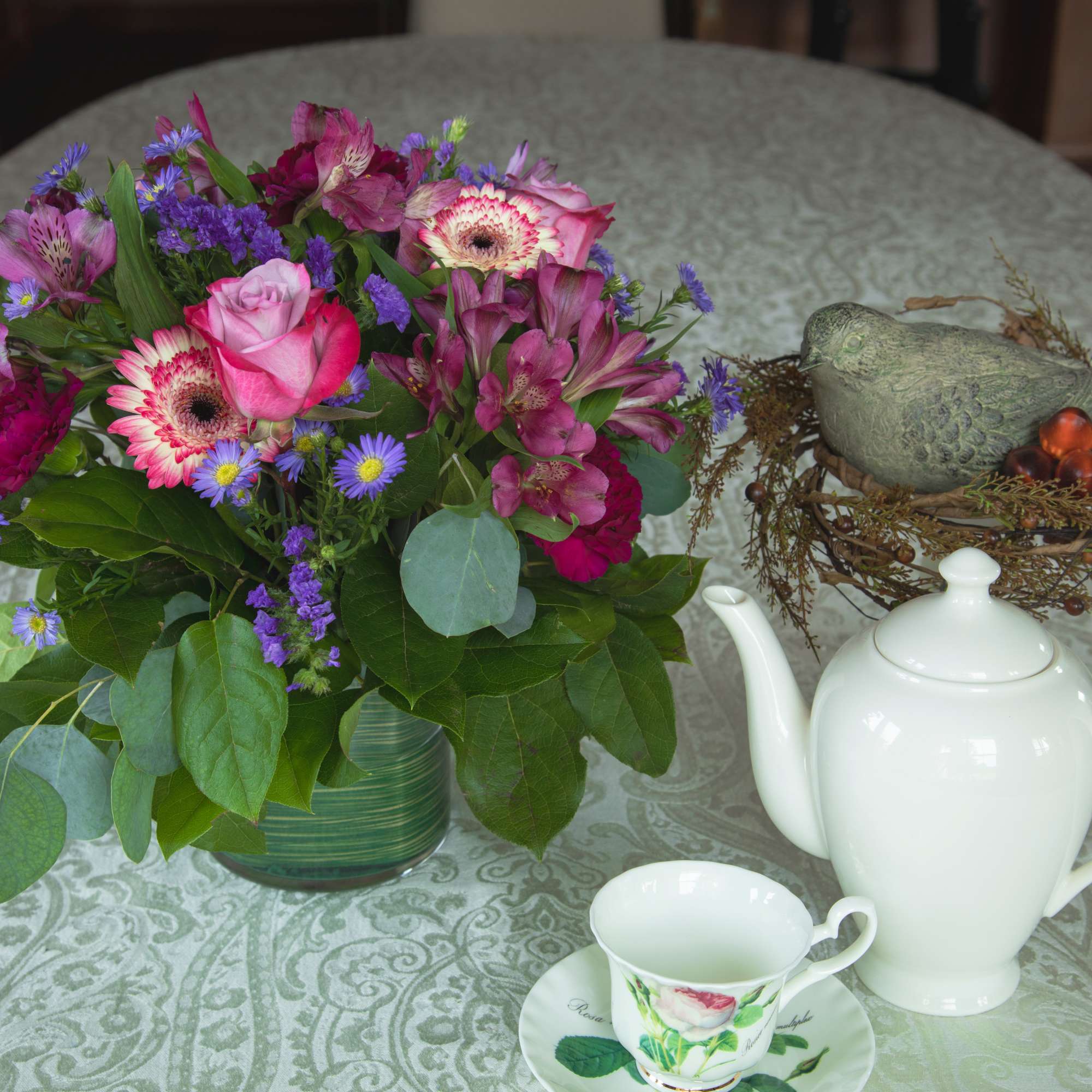 Pink and purple mixed bouquet in a glass vase beside a white teapot and cup