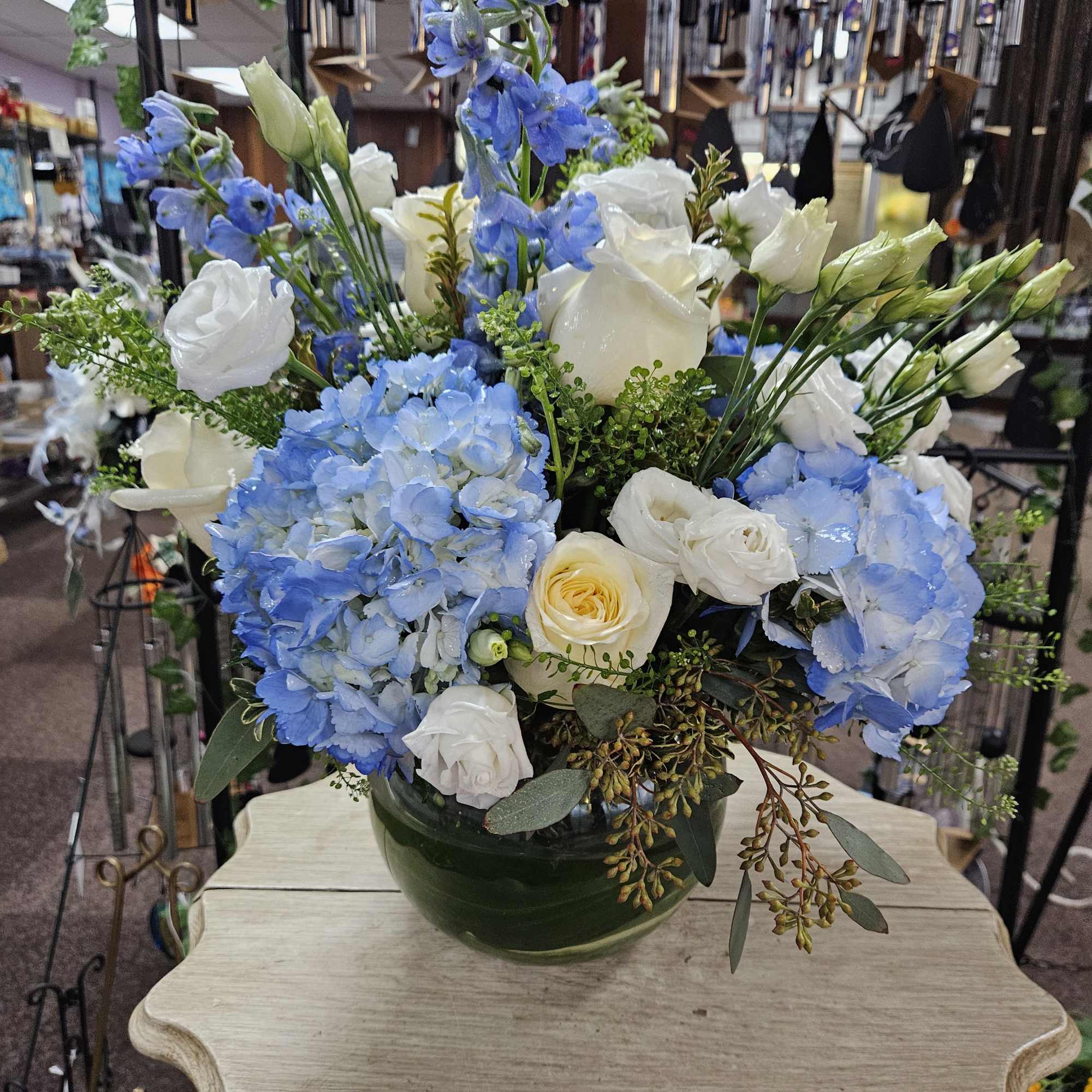Low glass bowl of blue hydrangeas, white and yellow roses with blue delphinium spikes.