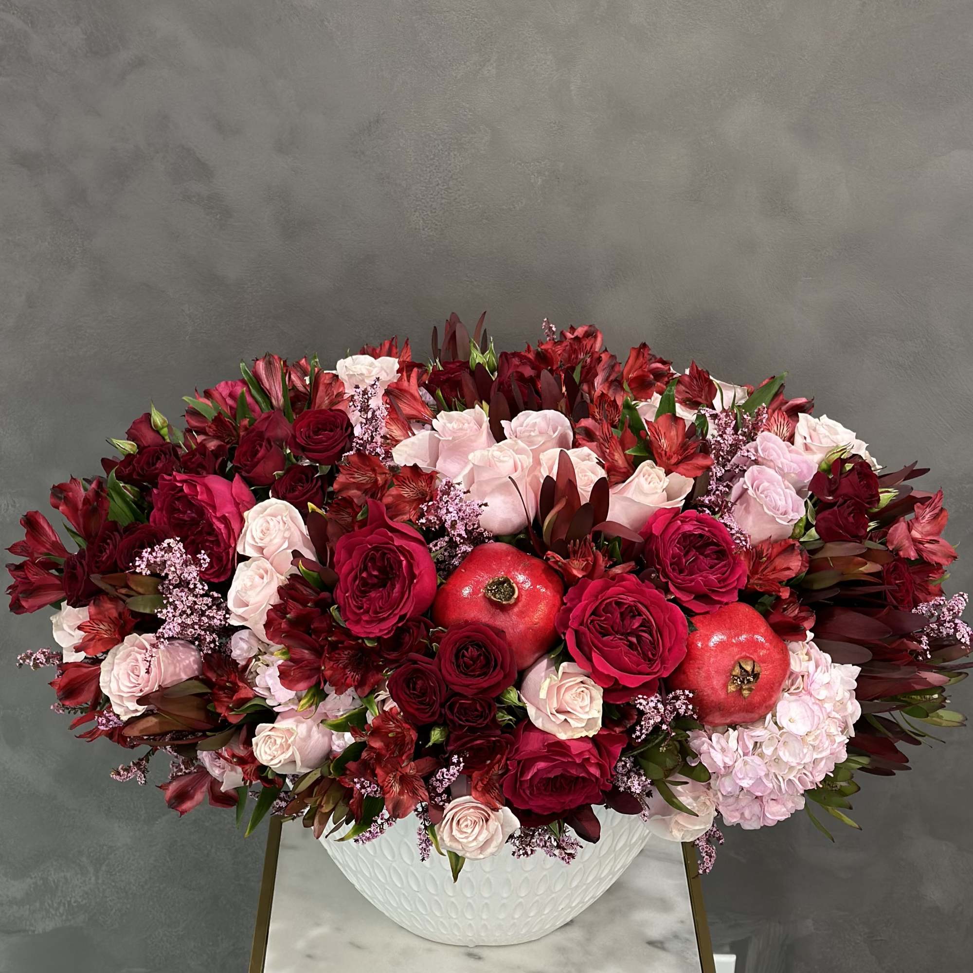 Large red and pink floral arrangement in a white bowl vase with pomegranates
