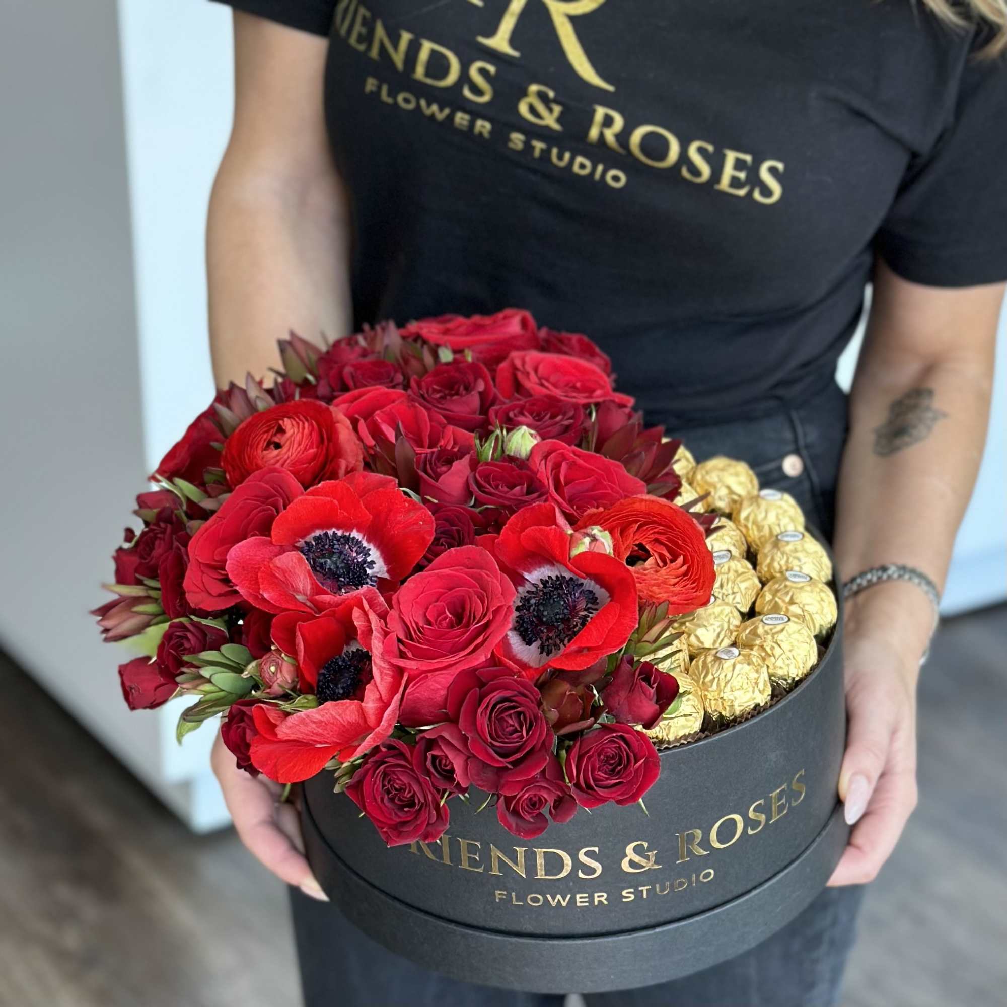 Red roses and poppies arranged in a round black box with gold chocolates.