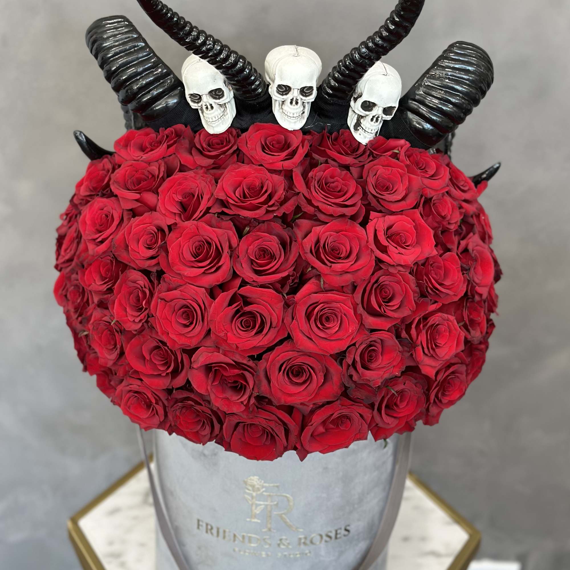 Red rose arrangement with black horns and skull decorations in a silver hatbox