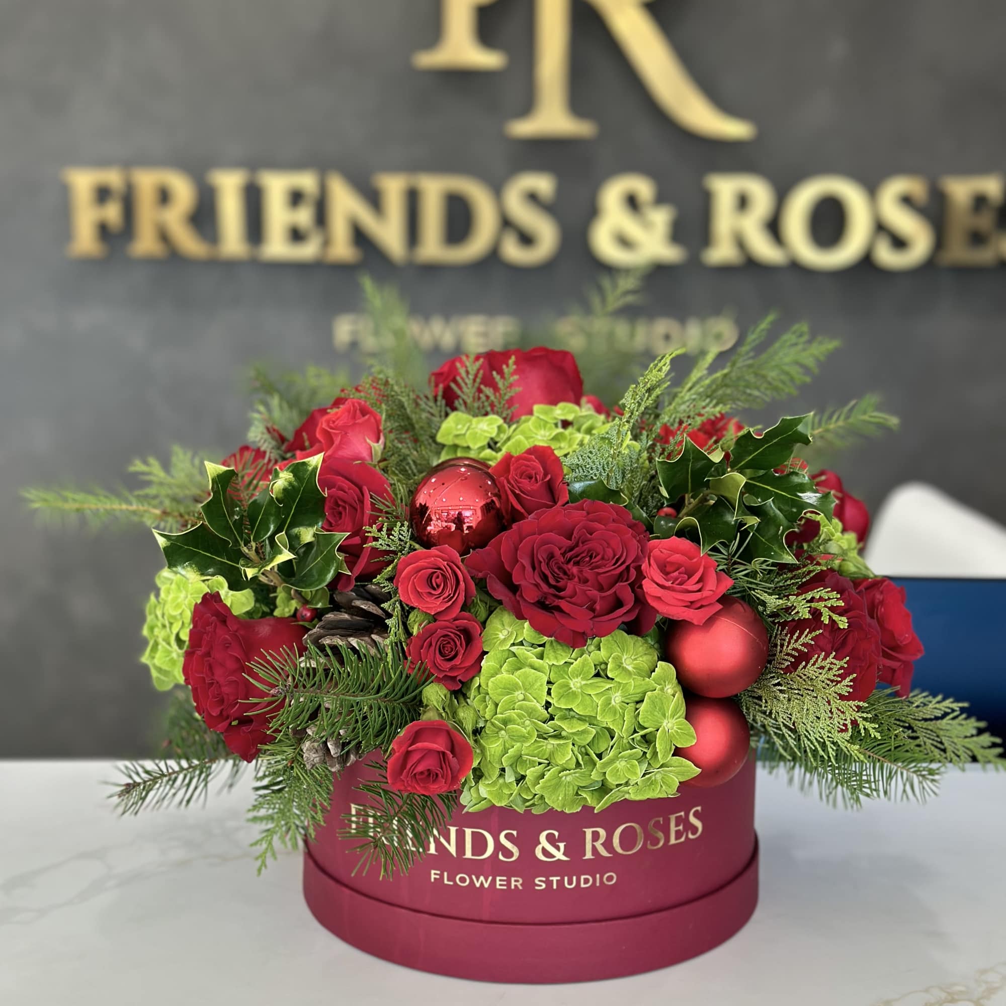 Red roses and green hydrangeas in a pink hat box with holiday ornaments.