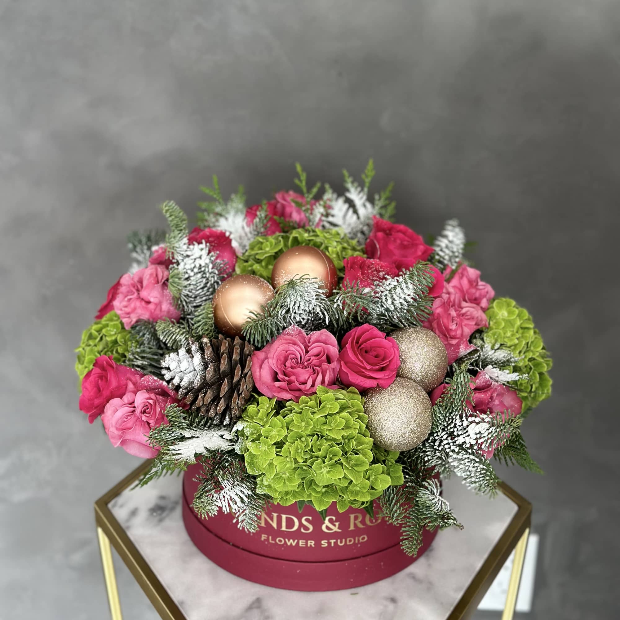 Pink roses and green hydrangeas arranged with pinecones and gold ornaments in a round box