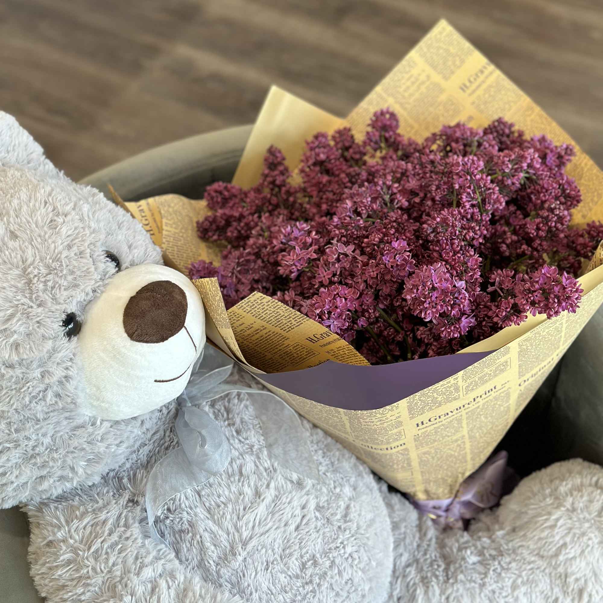 A teddy bear holding a bouquet of purple flowers wrapped in paper