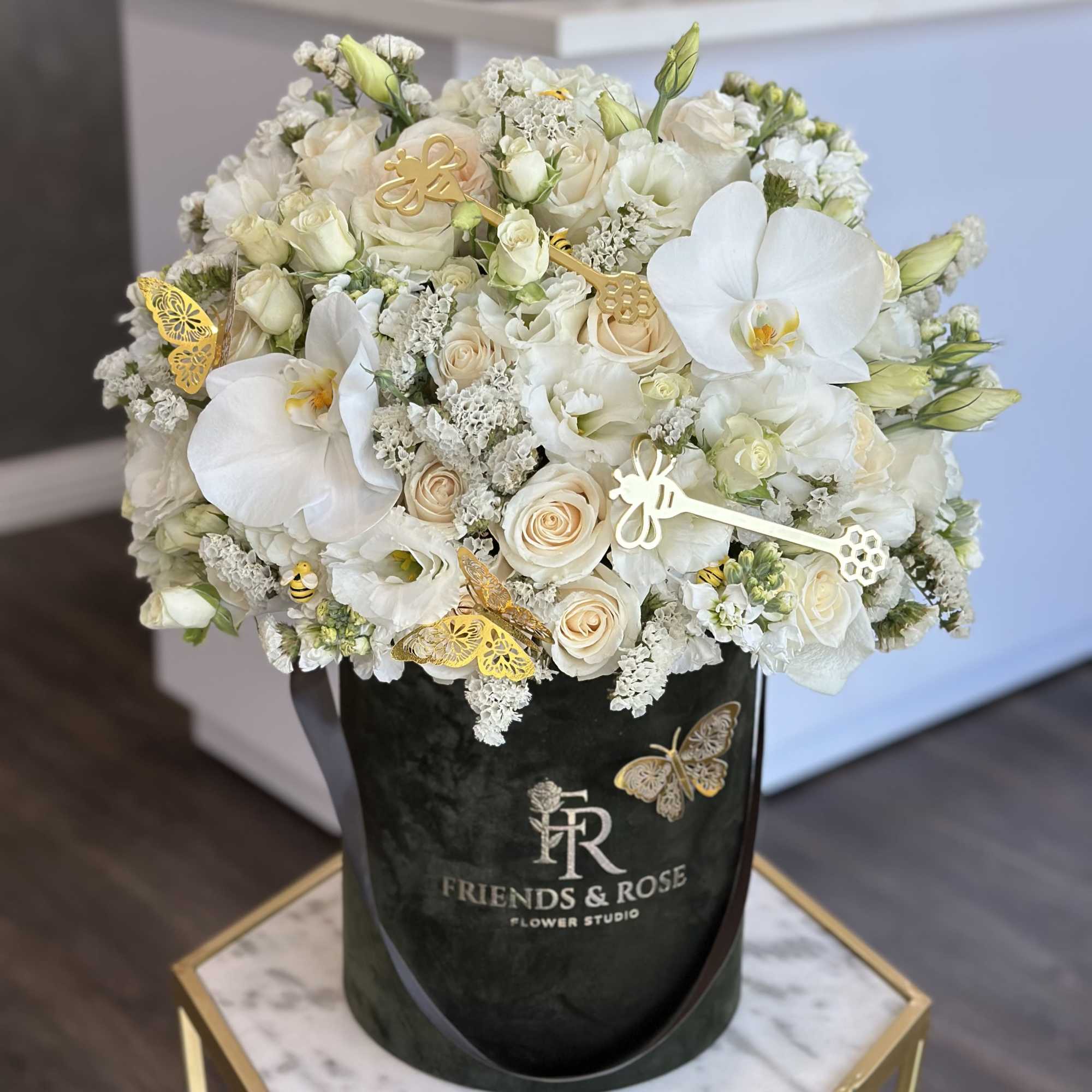 White roses and orchids arranged in a black hatbox with gold butterfly accents.