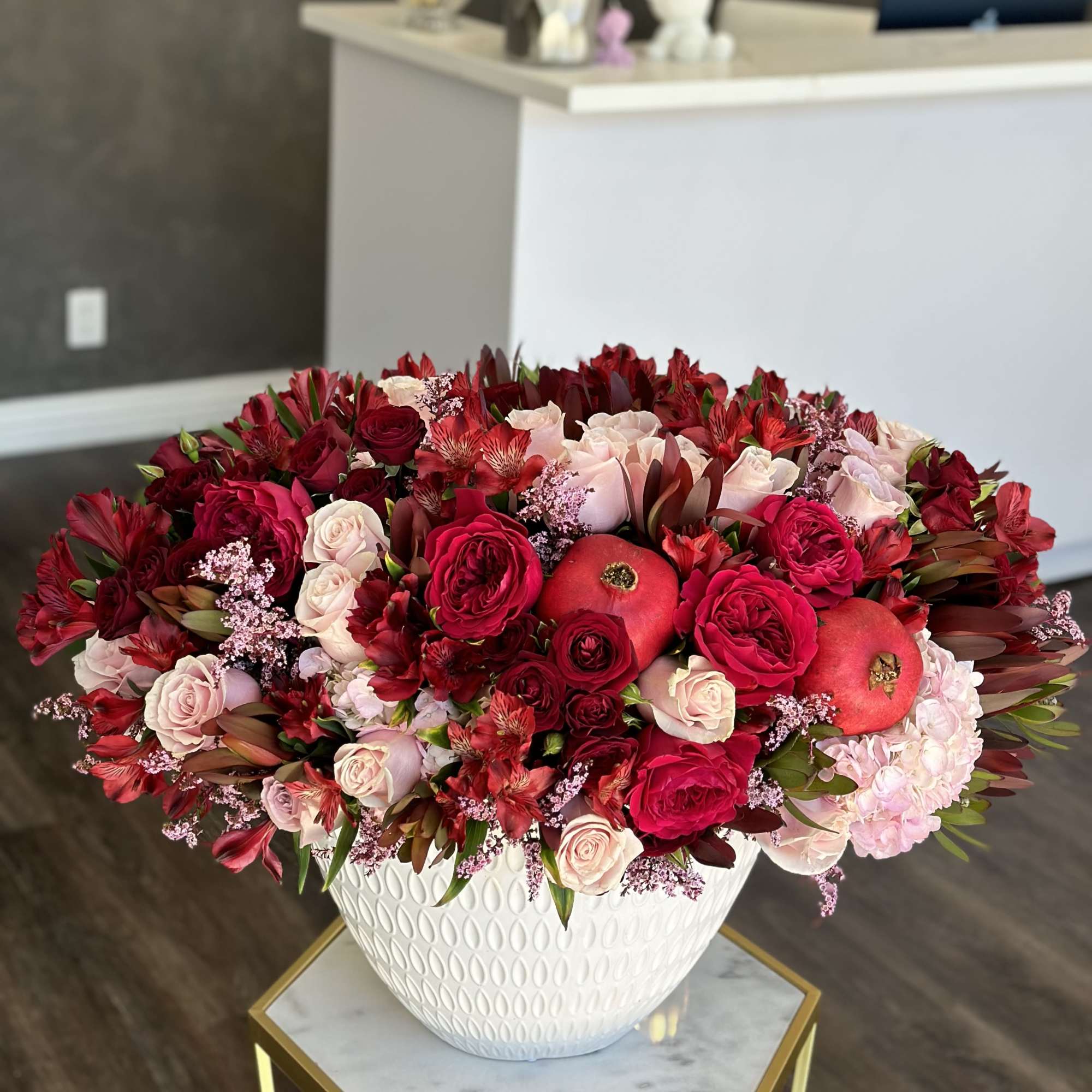 Large red and pink floral arrangement in a white vase with pomegranates