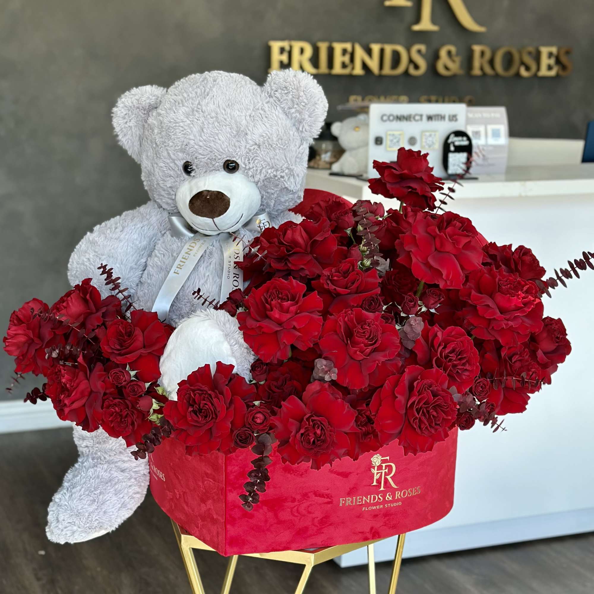Gray teddy bear beside a red rose arrangement in a pink box