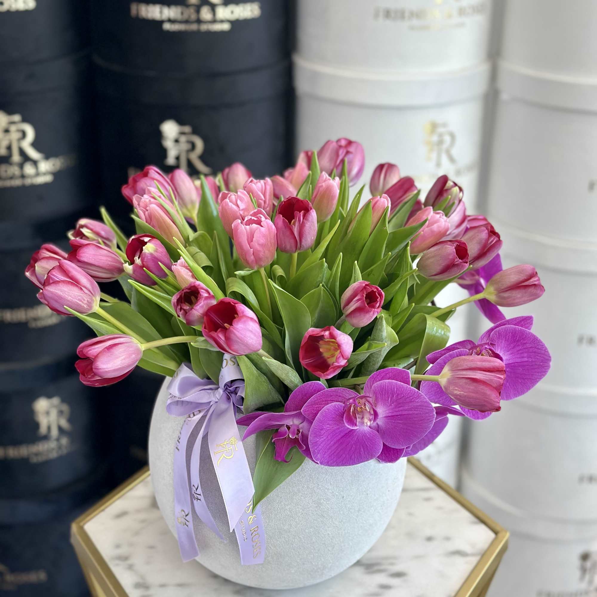 Pink tulips and purple orchids arranged in a round vase with a ribbon