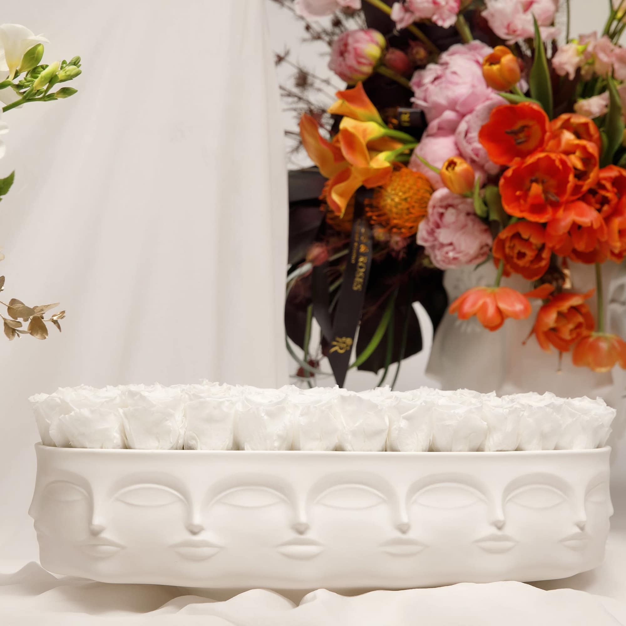 White roses in a low white rectangular vase with colorful bouquets behind