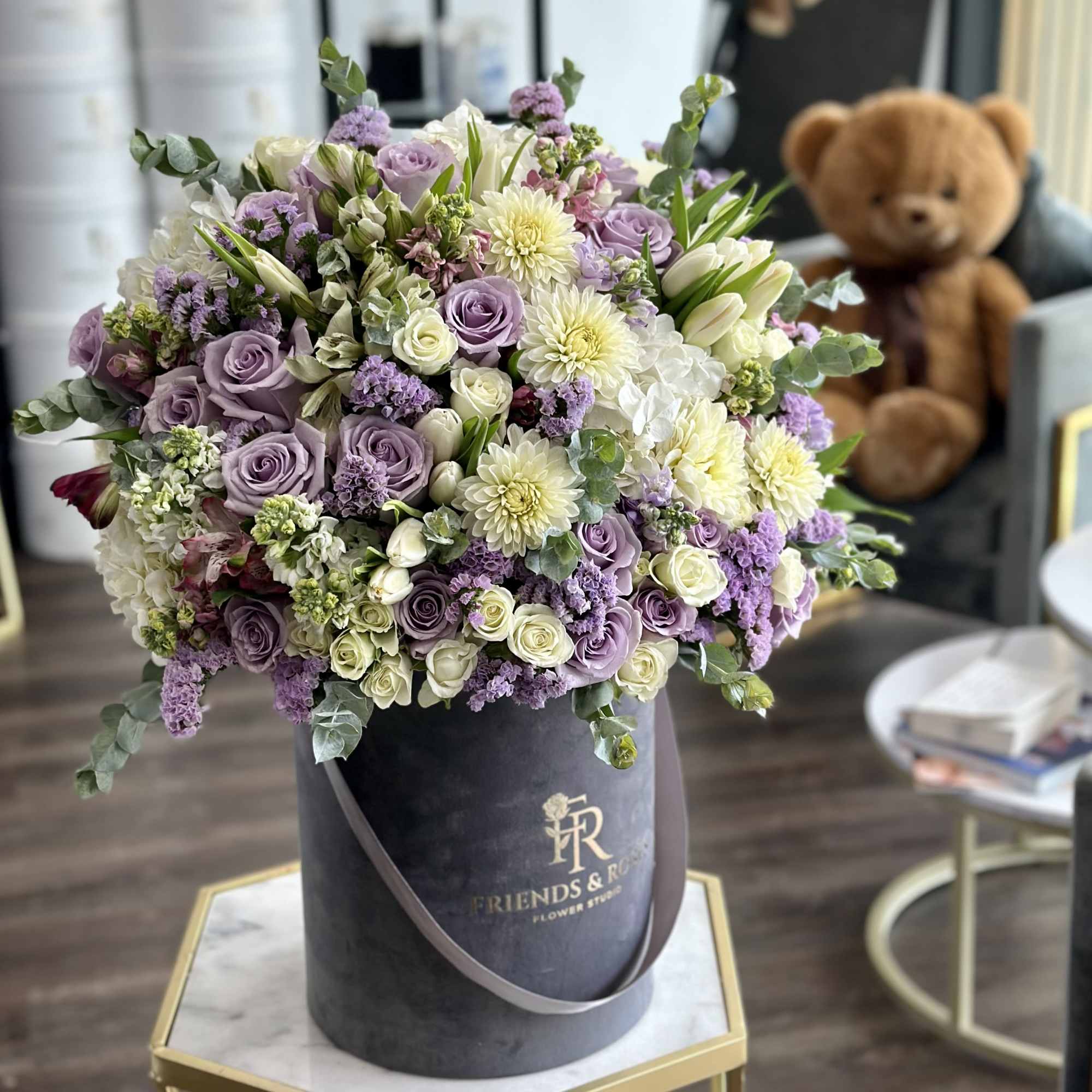 Lavender and white rose bouquet in a black hatbox with a teddy bear behind it