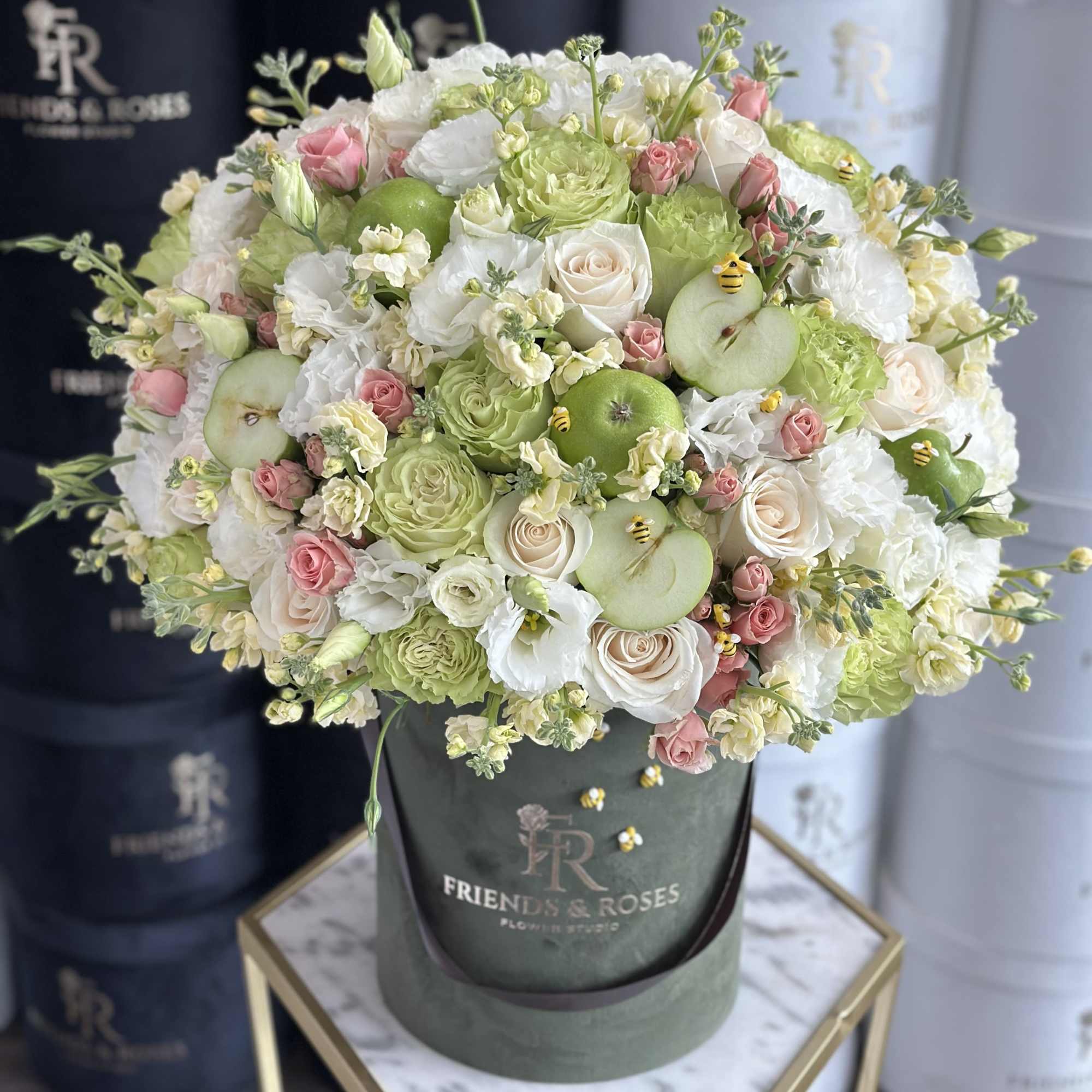 Round bouquet of white, pink, and green flowers in a green hatbox with apple accents.