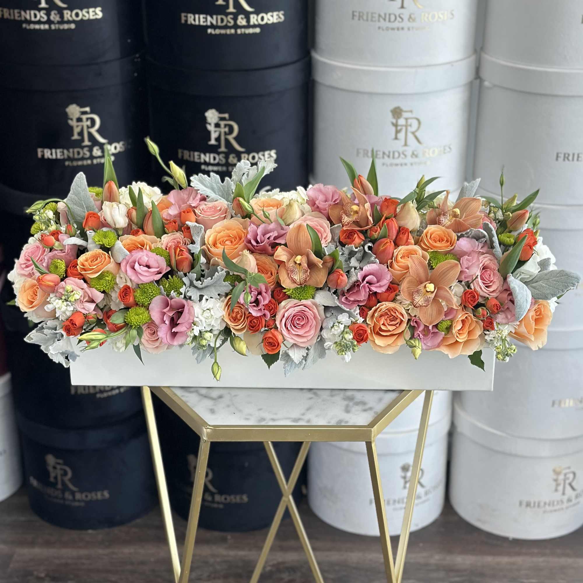 Low rectangular box arrangement of peach, orange, and pink roses with orchids and mixed blooms.