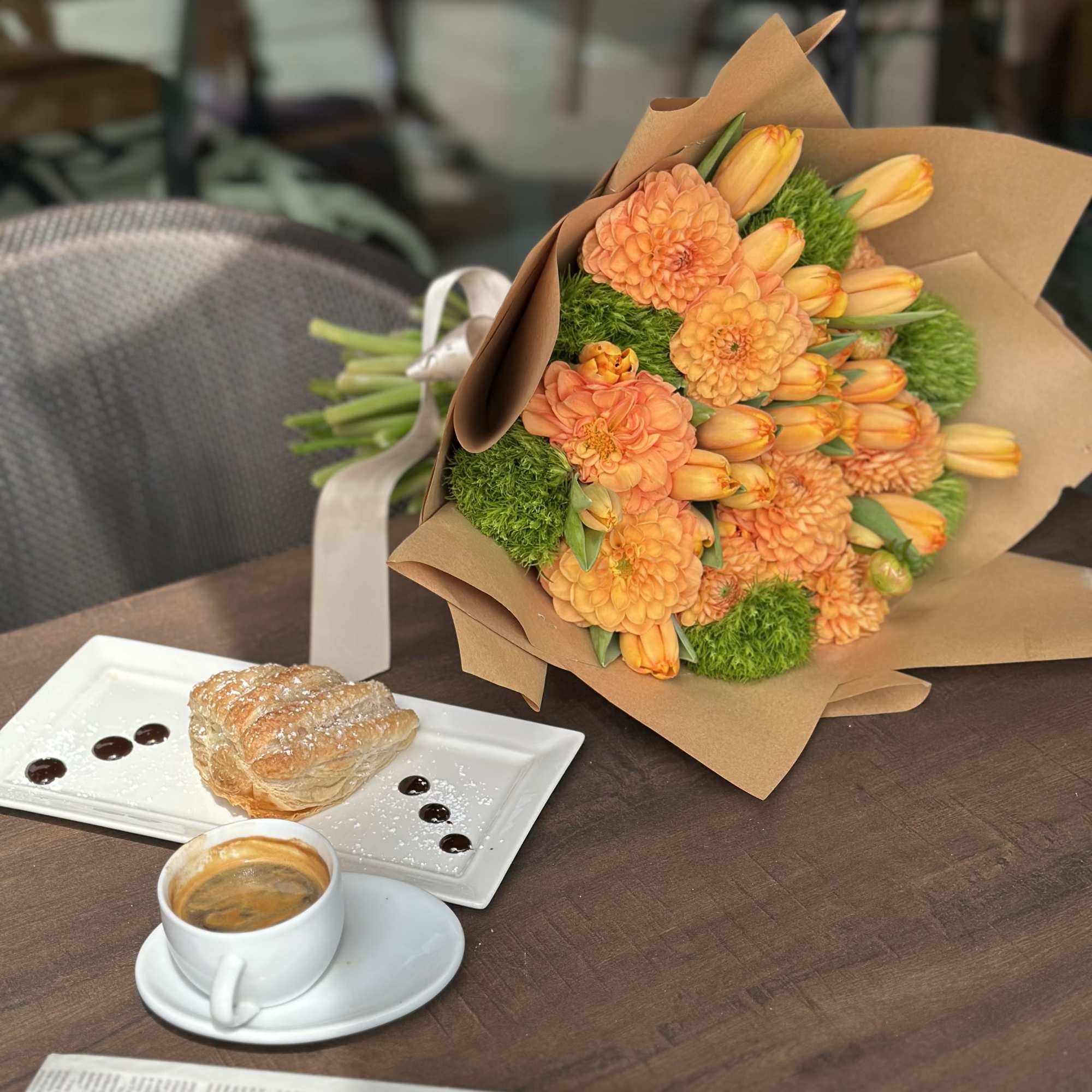 Orange flower bouquet wrapped in brown paper beside coffee and pastry