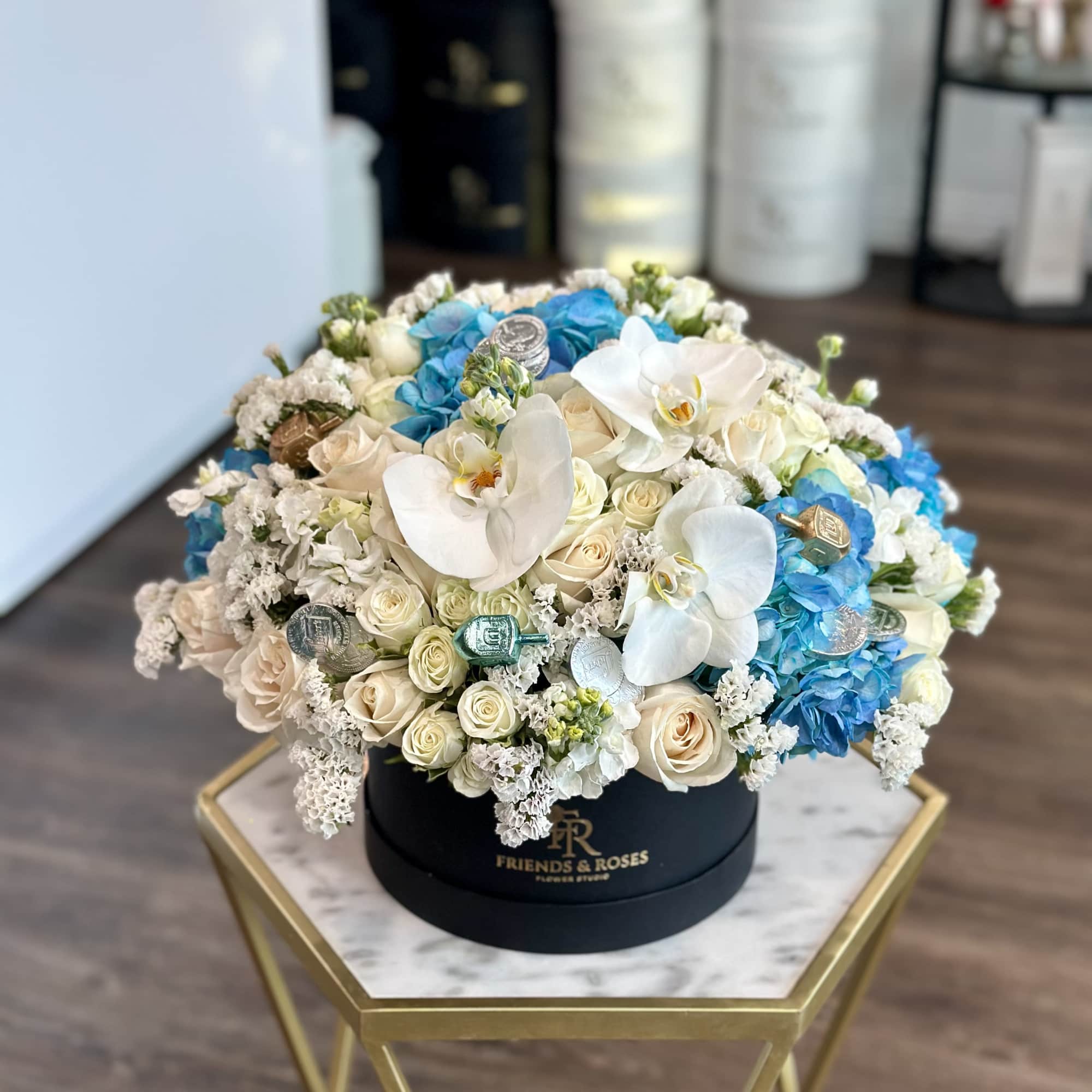 Round floral arrangement with white orchids, cream roses, and blue hydrangeas in a black box.