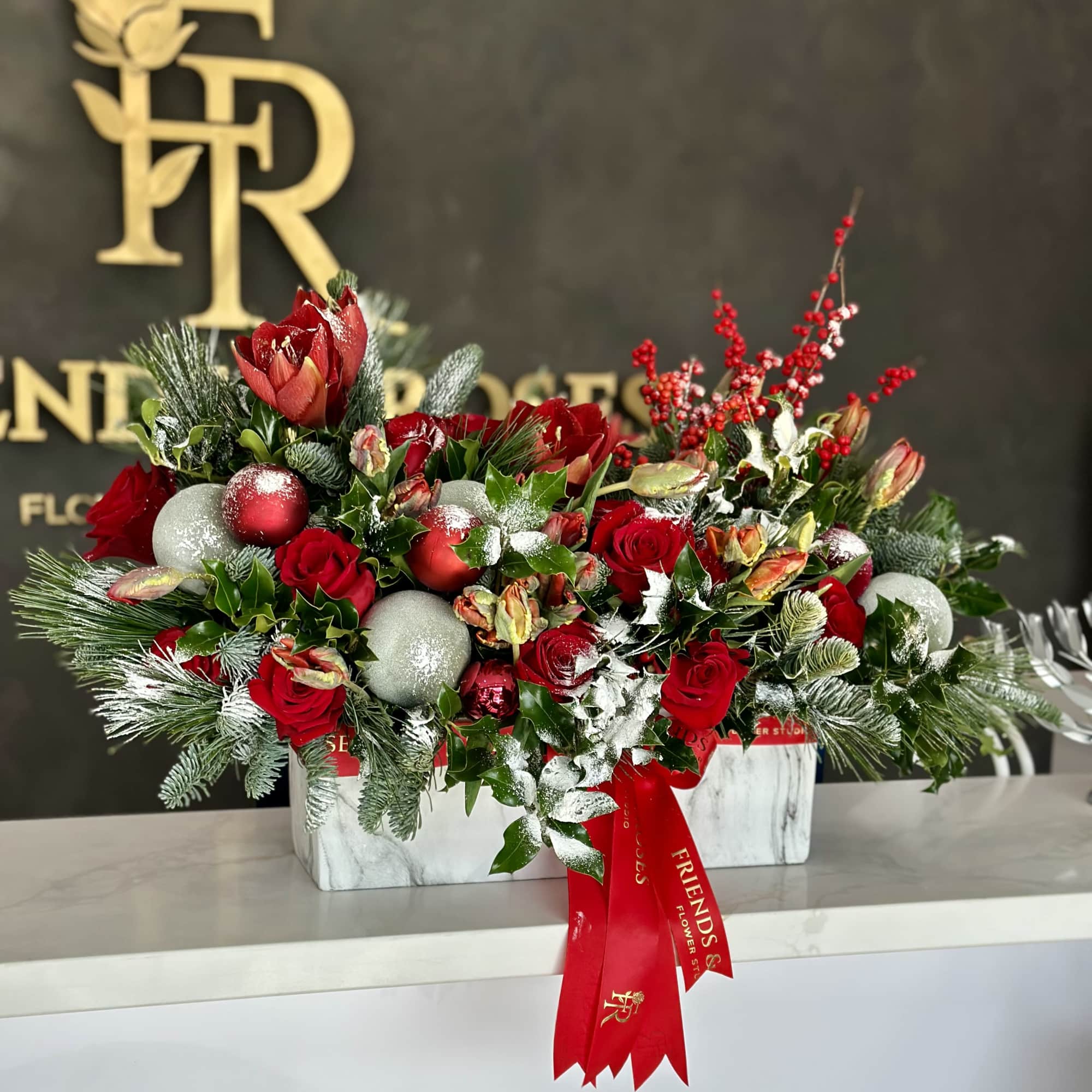 Red roses and holiday greenery arranged in a white rectangular box with ornaments.