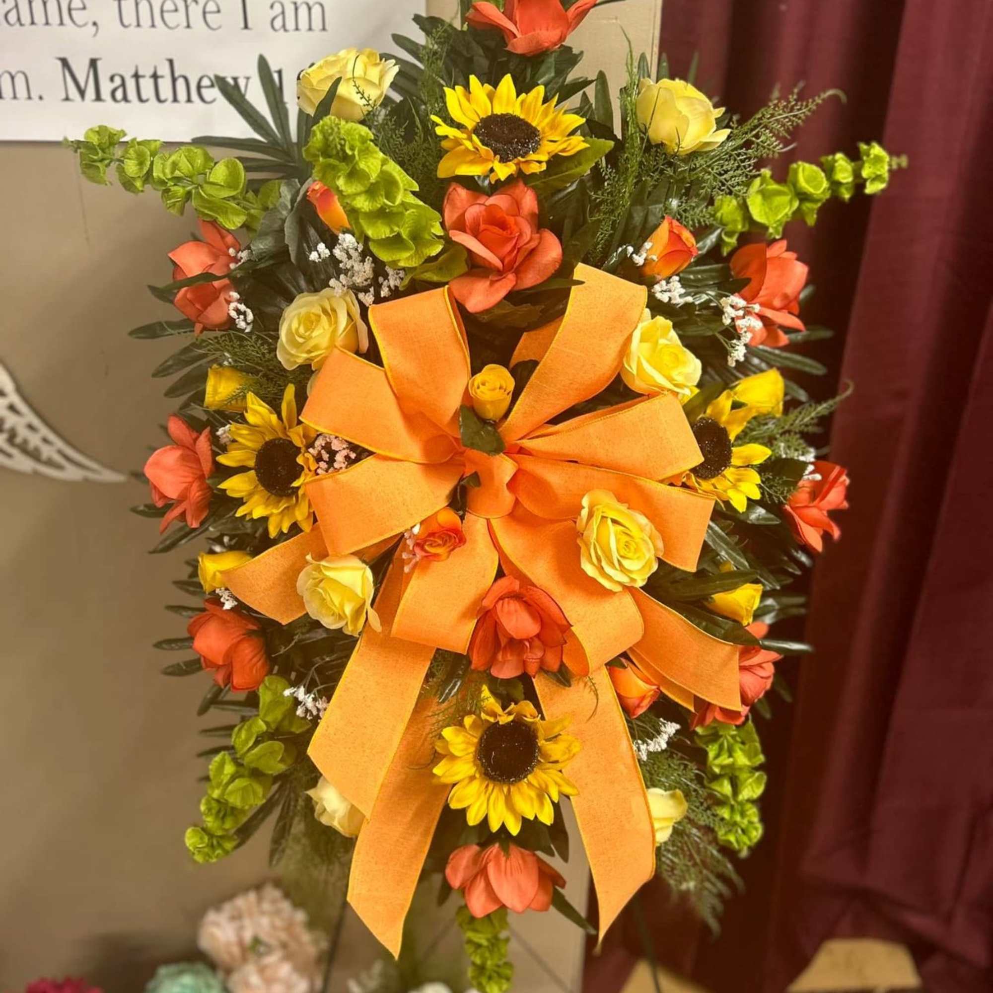 Orange ribbon wreath with yellow sunflowers and roses on a stand