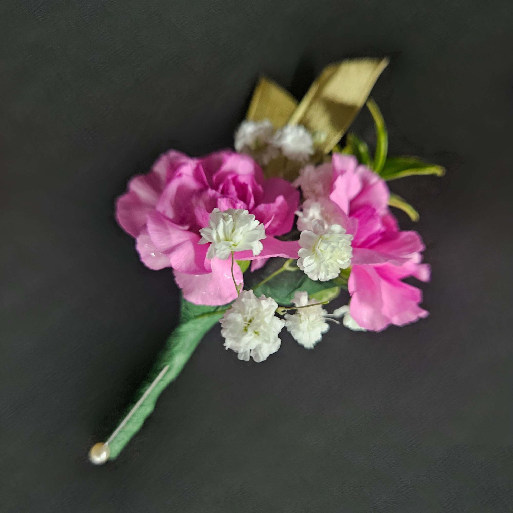 Pink carnation and white filler flower boutonniere with gold ribbon and pearl pin on a dark background