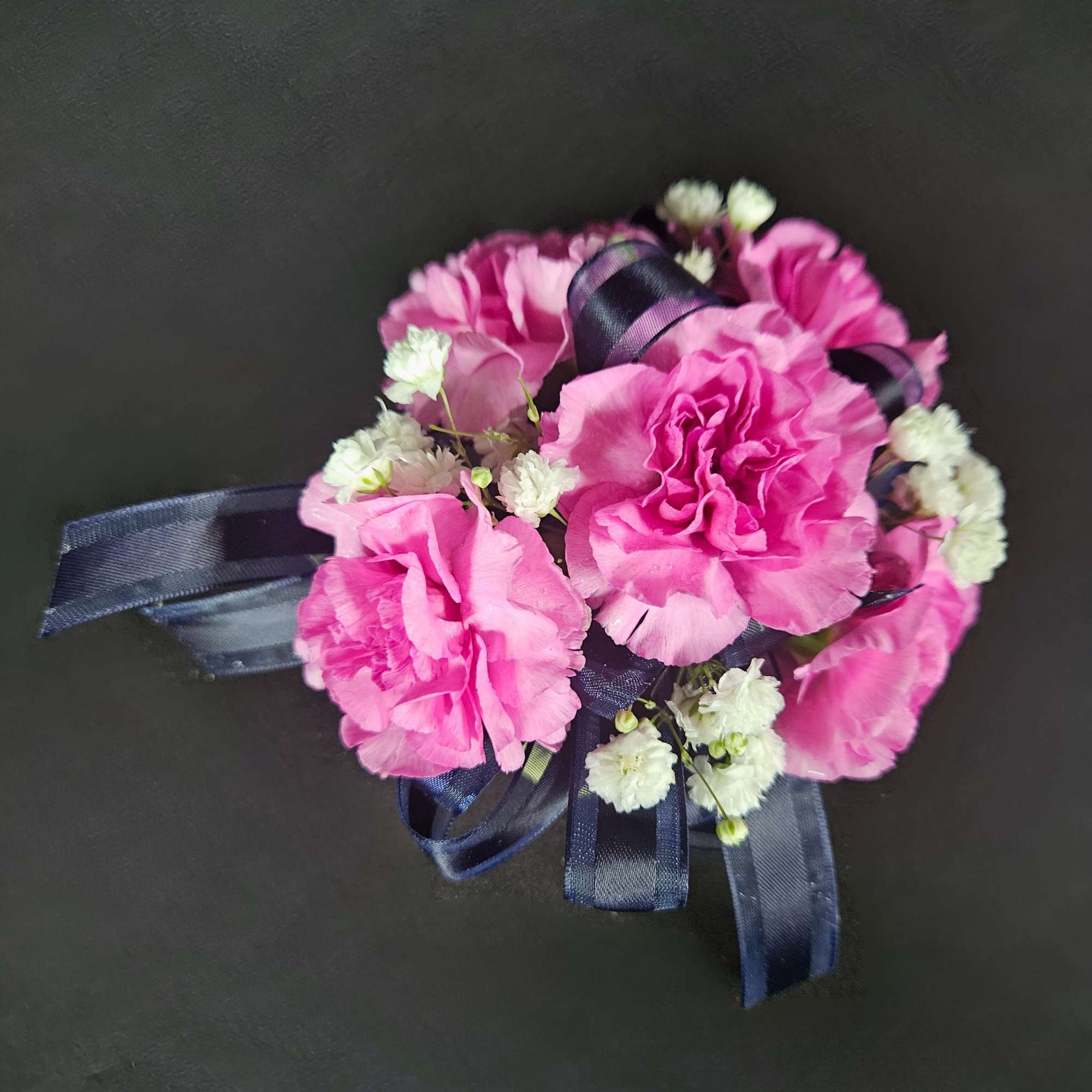 Pink carnation corsage with white filler flowers and navy ribbon on a dark background