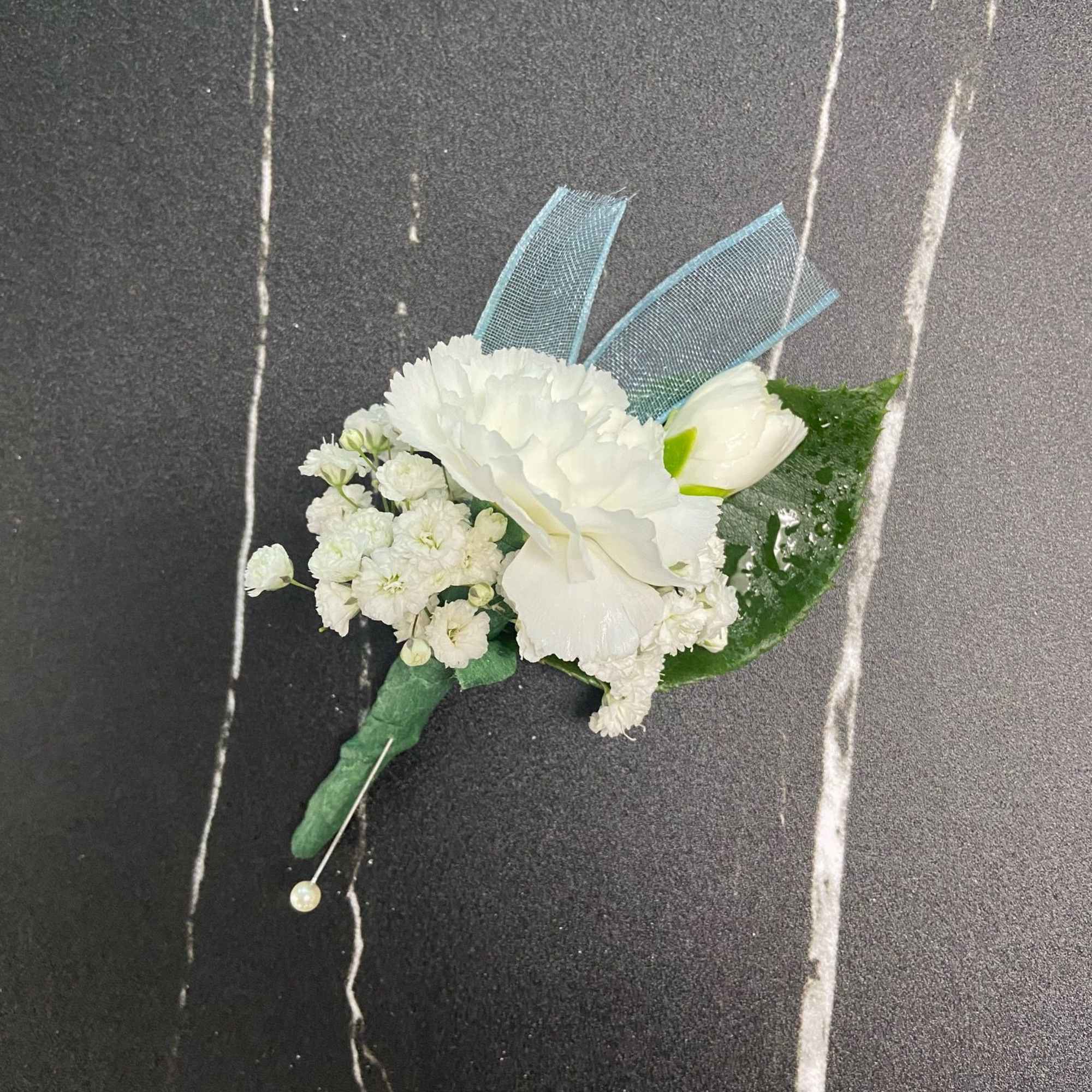 White carnation boutonniere with small white blooms, blue ribbon, and pearl pin on a dark background