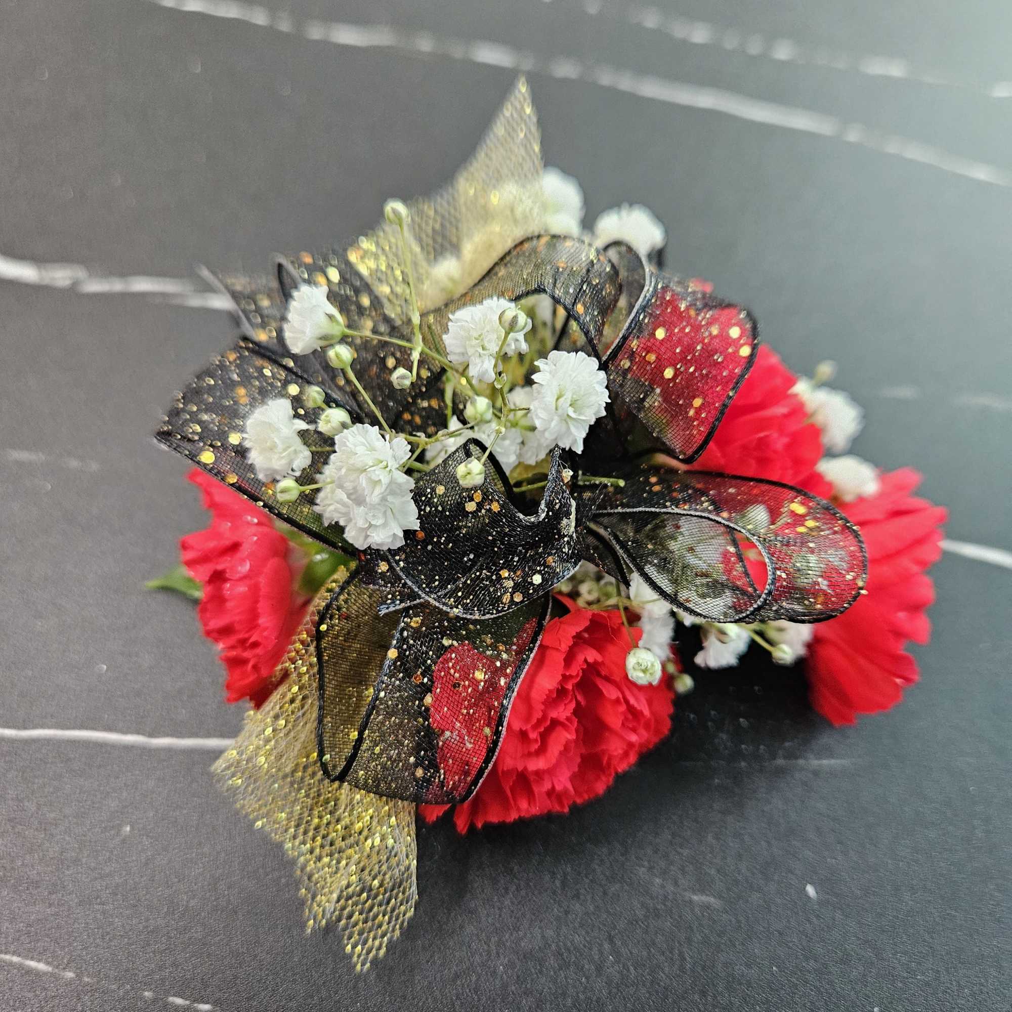 Red carnation corsage with white filler flowers and black and gold glitter ribbon on a dark surface