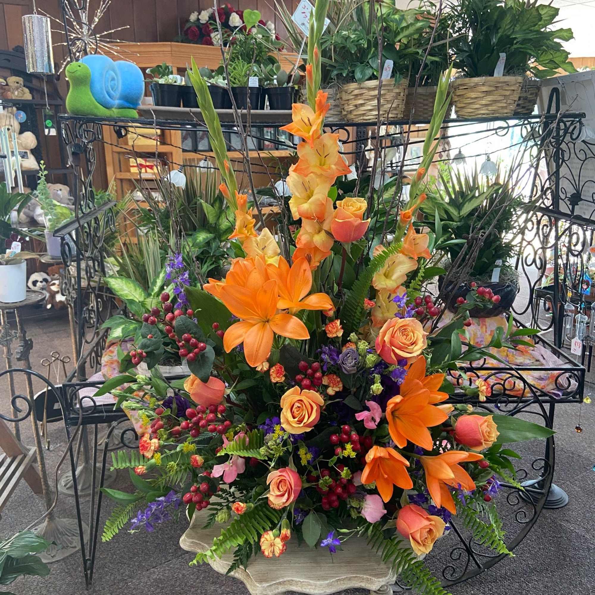 Tall arrangement of orange lilies, roses, and gladiolus with mixed blooms in a lush container.