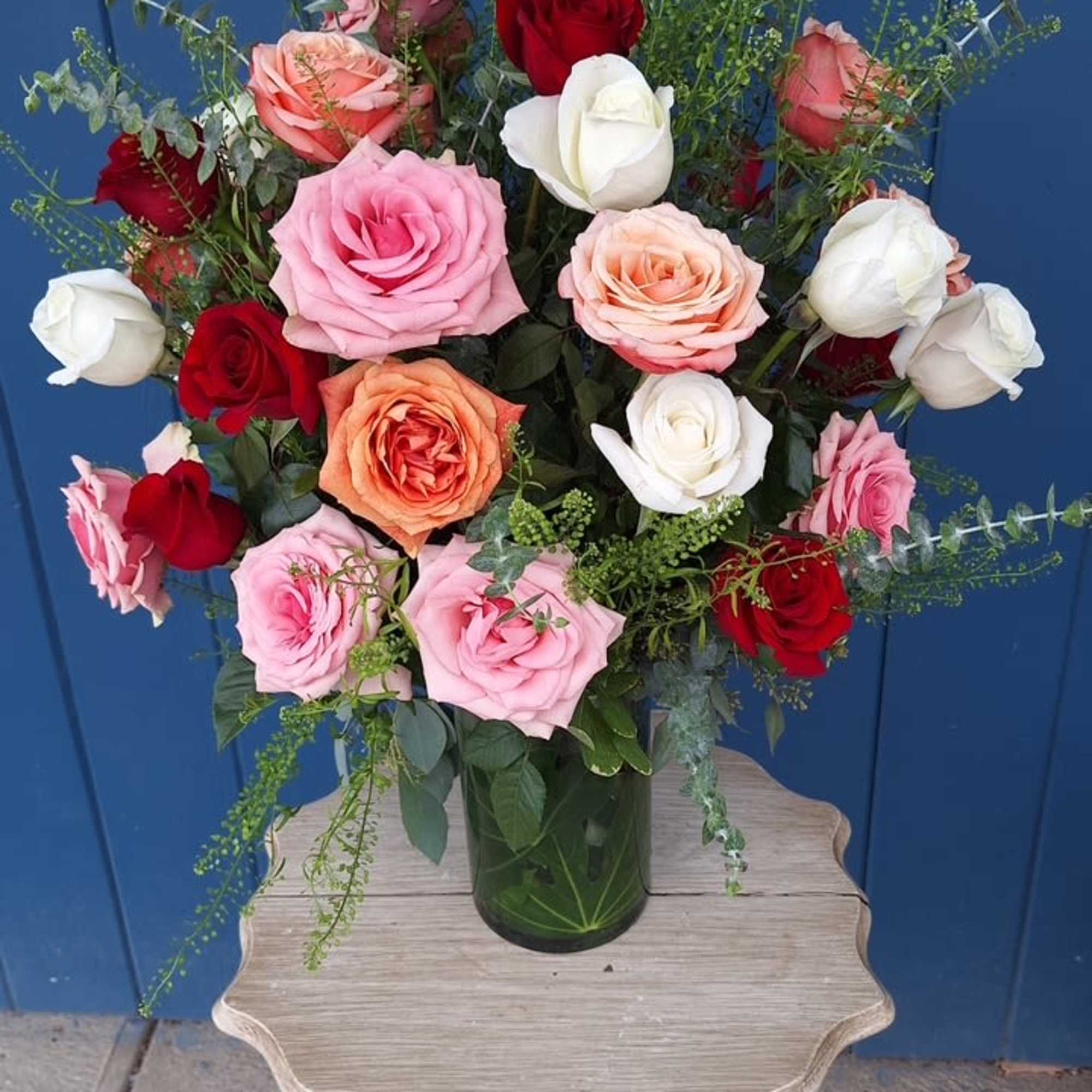Tall mixed rose arrangement in a clear vase with greenery on a small wooden table.