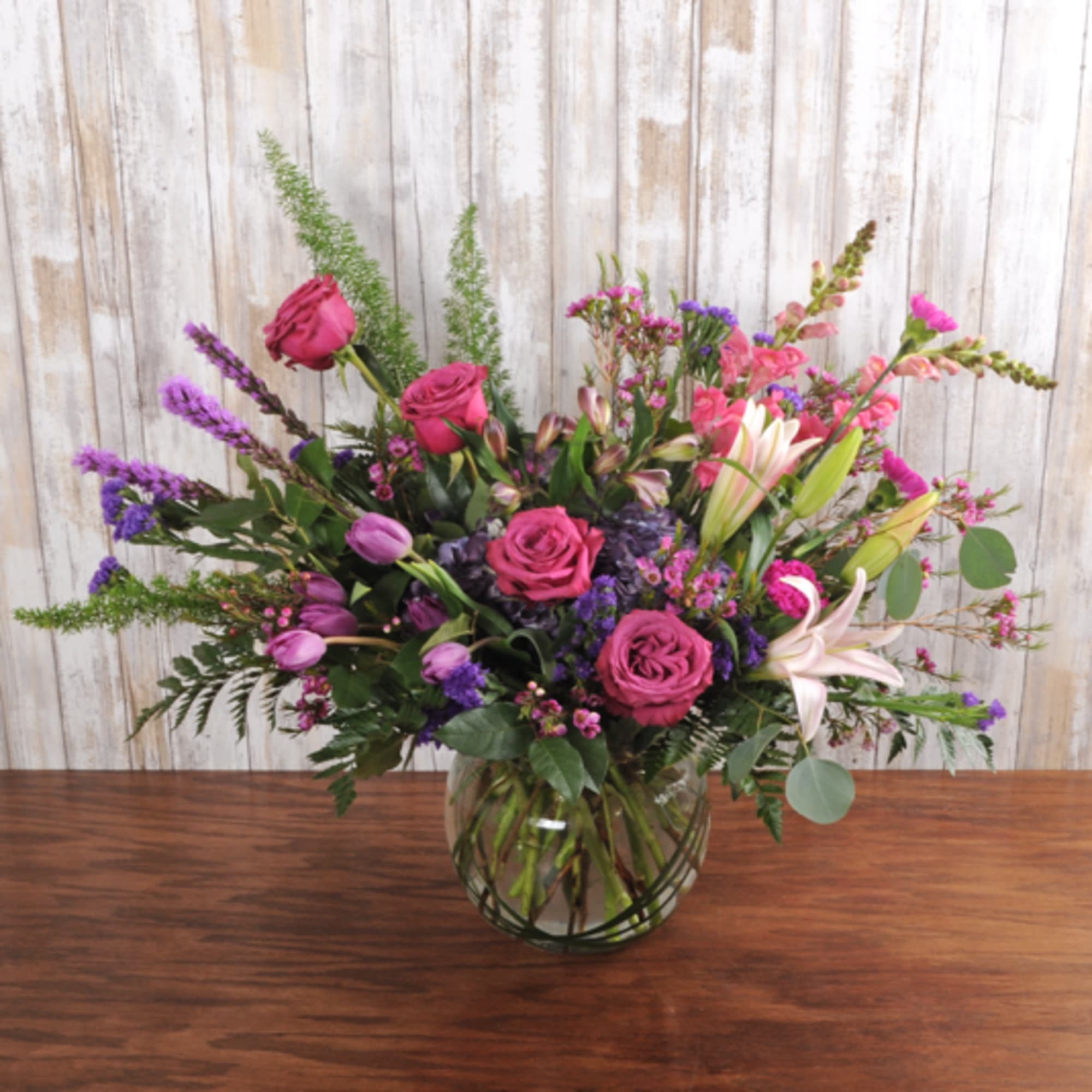 Large mixed bouquet of pink roses, lilies and purple blooms in a round clear glass vase on a wooden table