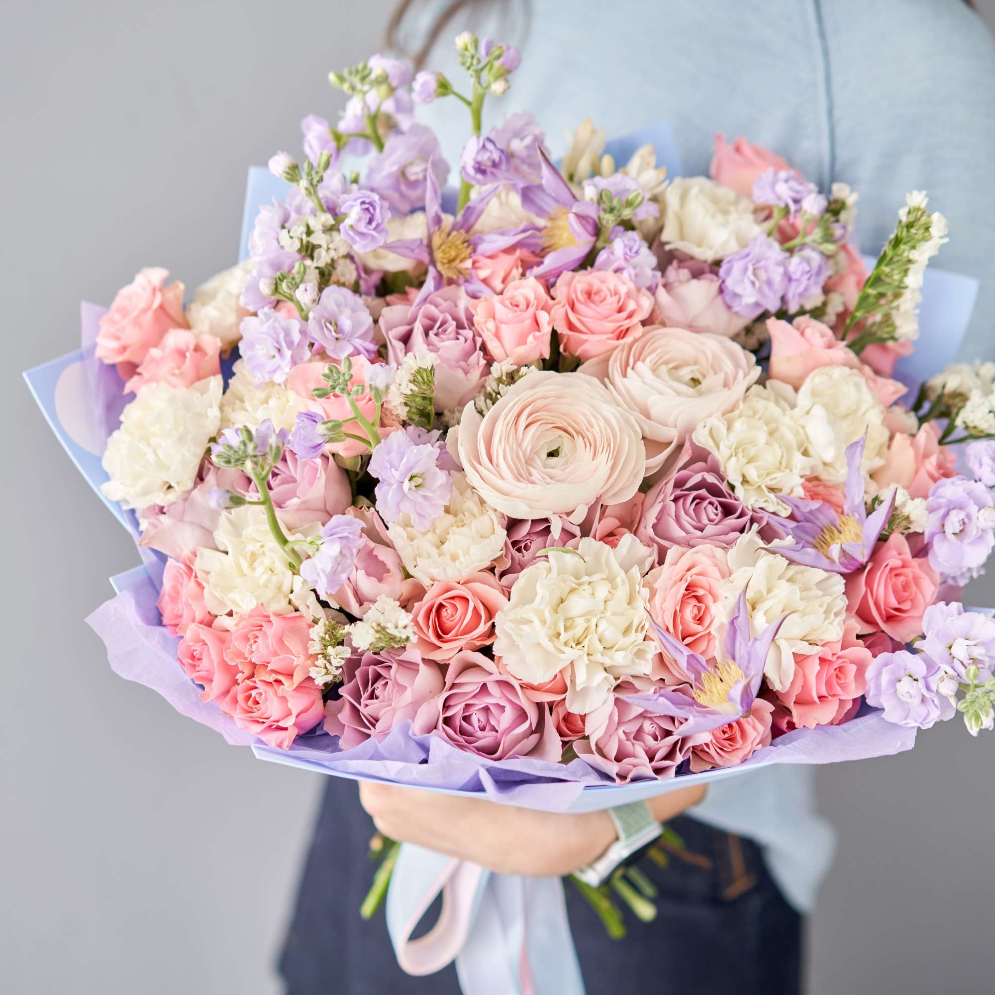 Large pastel bouquet of pink roses, cream blooms, and lavender flowers wrapped in purple paper.
