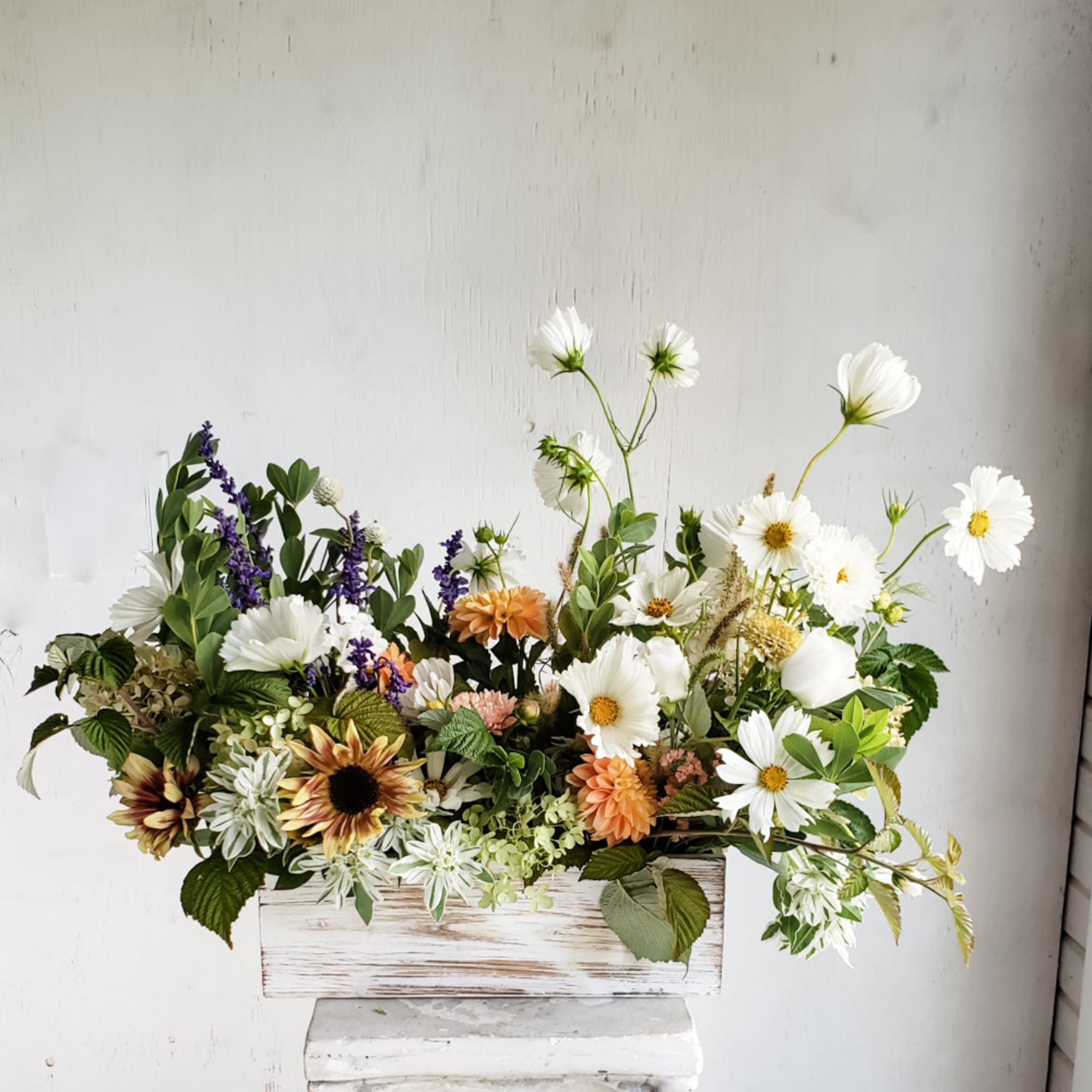 Mixed bouquet of white daisies, orange blooms, and purple flowers in a white wooden box