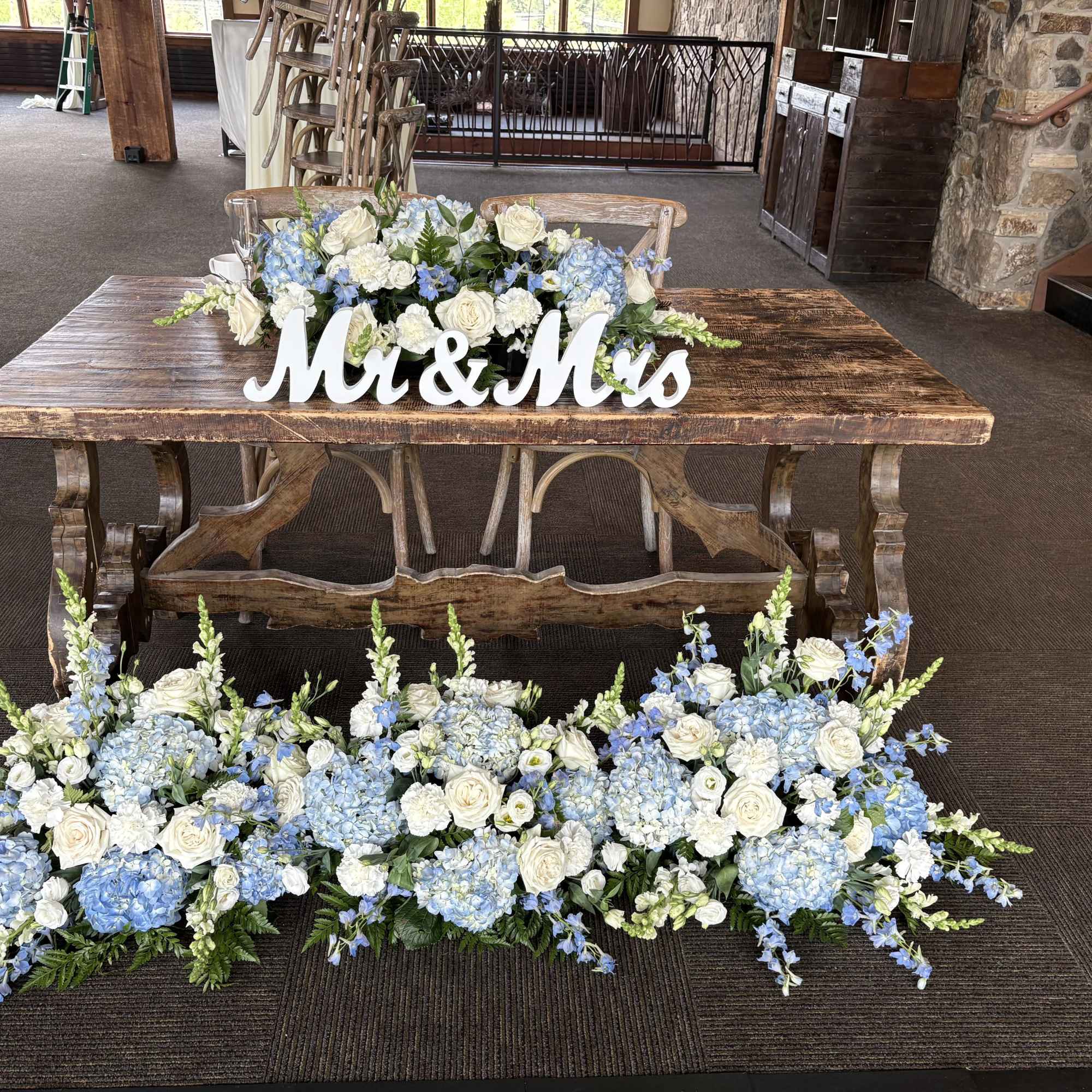 Rustic sweetheart table with blue and white floral centerpieces and Mr & Mrs sign, with matching floor arrangements