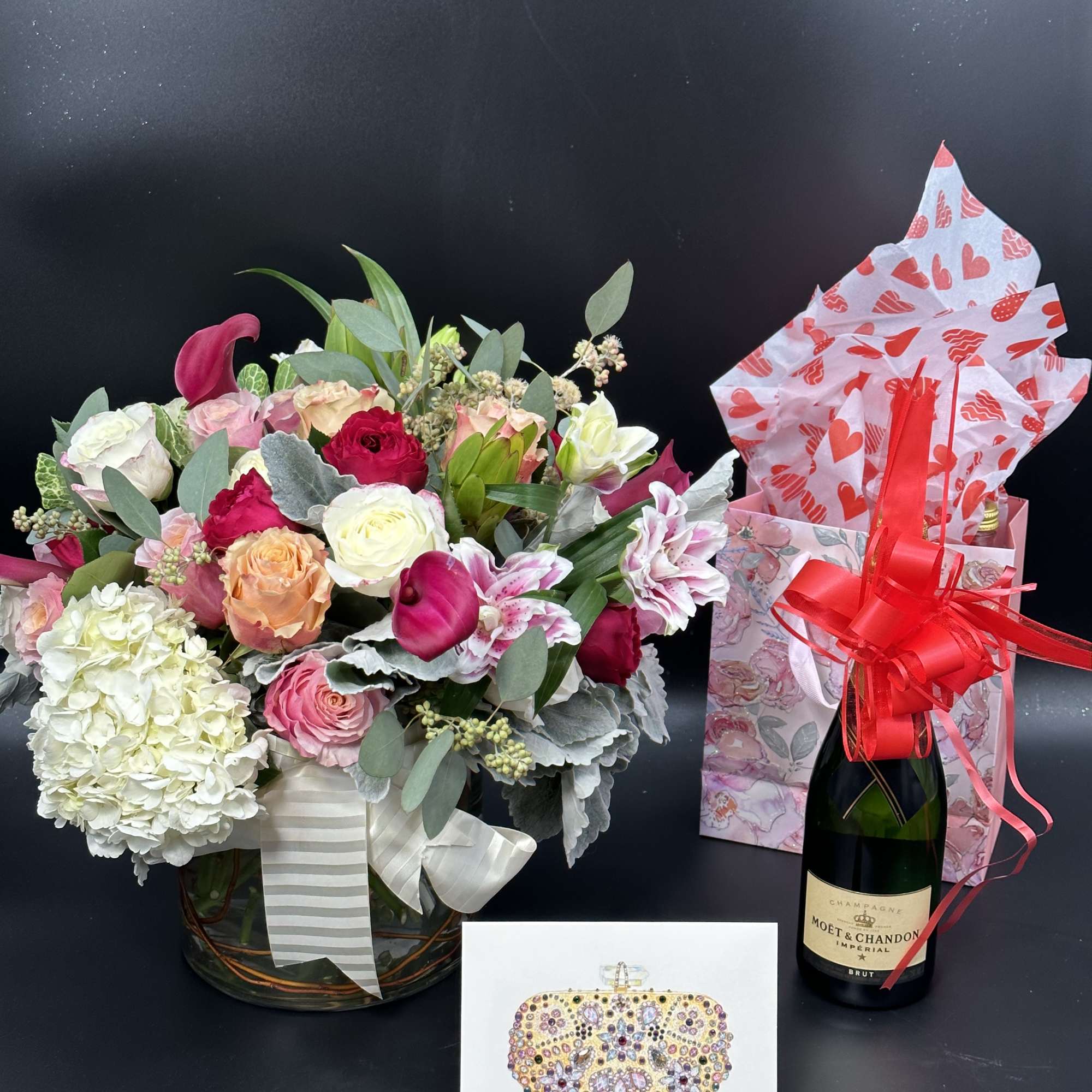Mixed flower arrangement in a glass vase with a champagne bottle and gift bag