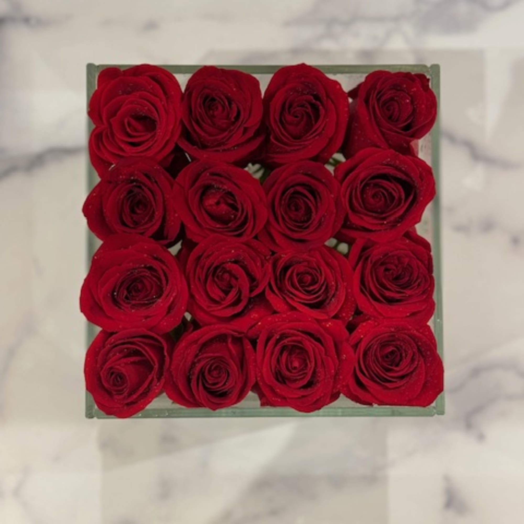 Square arrangement of red roses tightly packed in a clear container on a marble surface