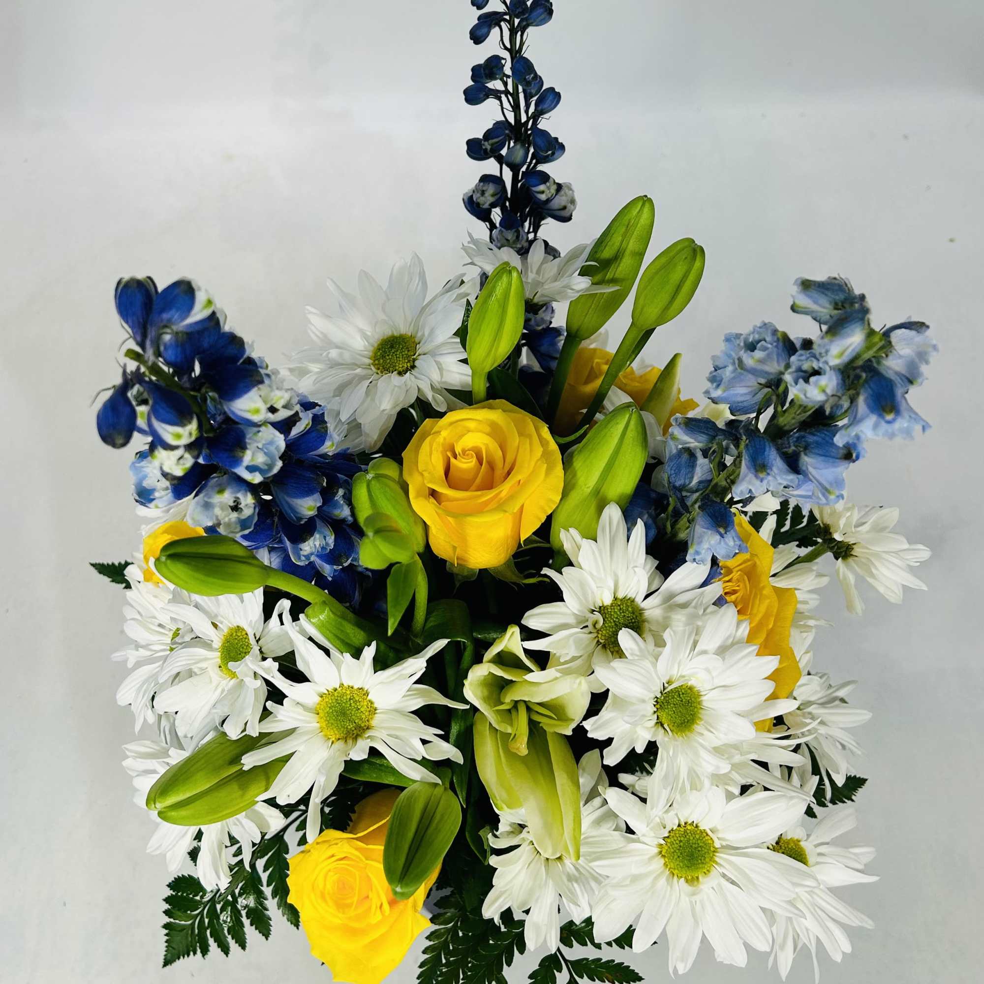 Yellow roses, white daisies, blue delphinium, and lilies in a bouquet