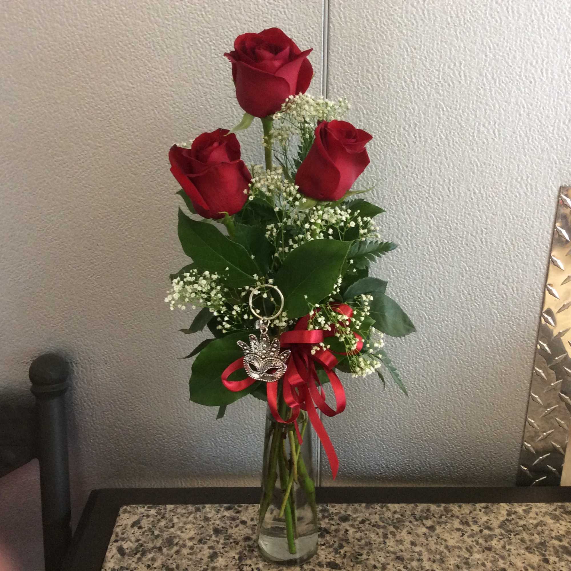 Three red roses in a clear glass vase with a red ribbon and charm