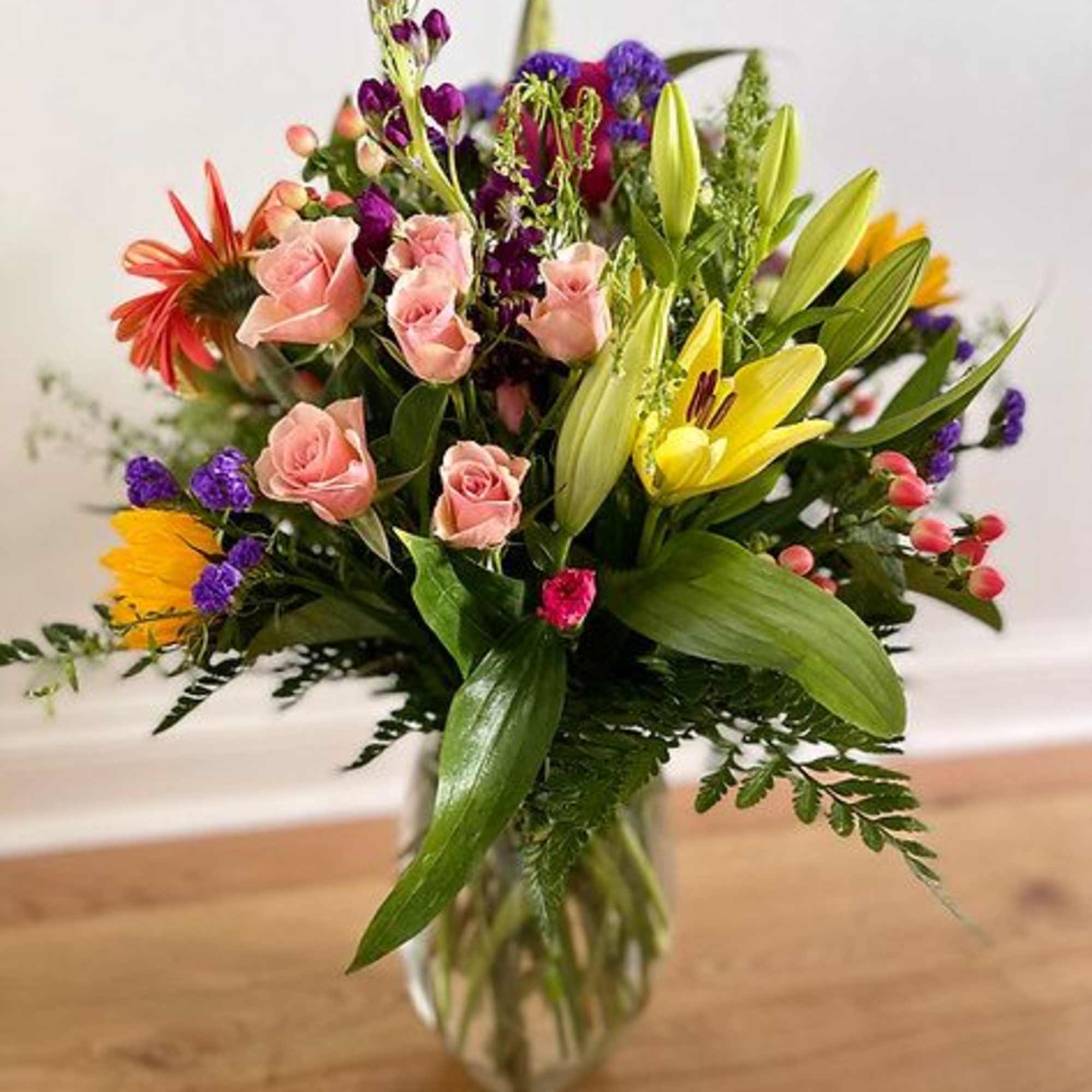 Mixed bouquet of pink roses, yellow lilies, and purple flowers in a glass vase