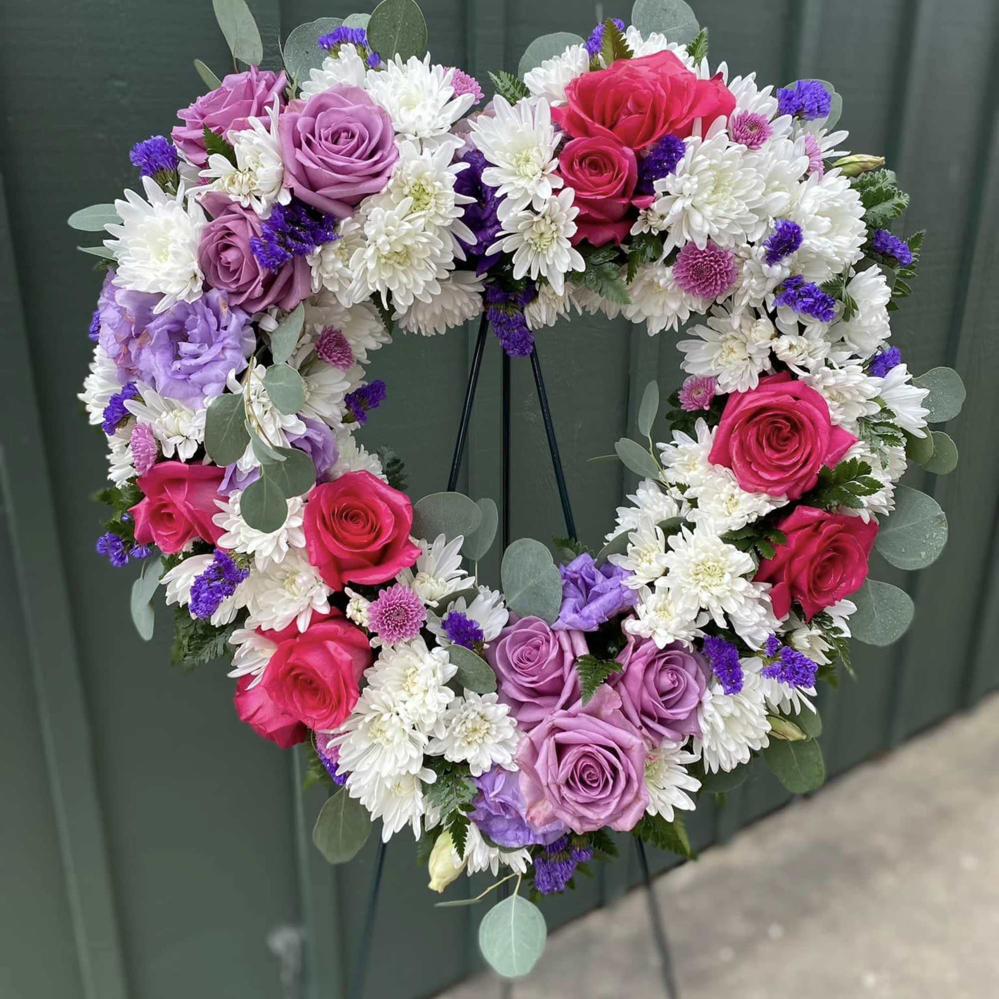 Heart-shaped wreath of pink and lavender roses with white mums and purple accents on a standing easel.