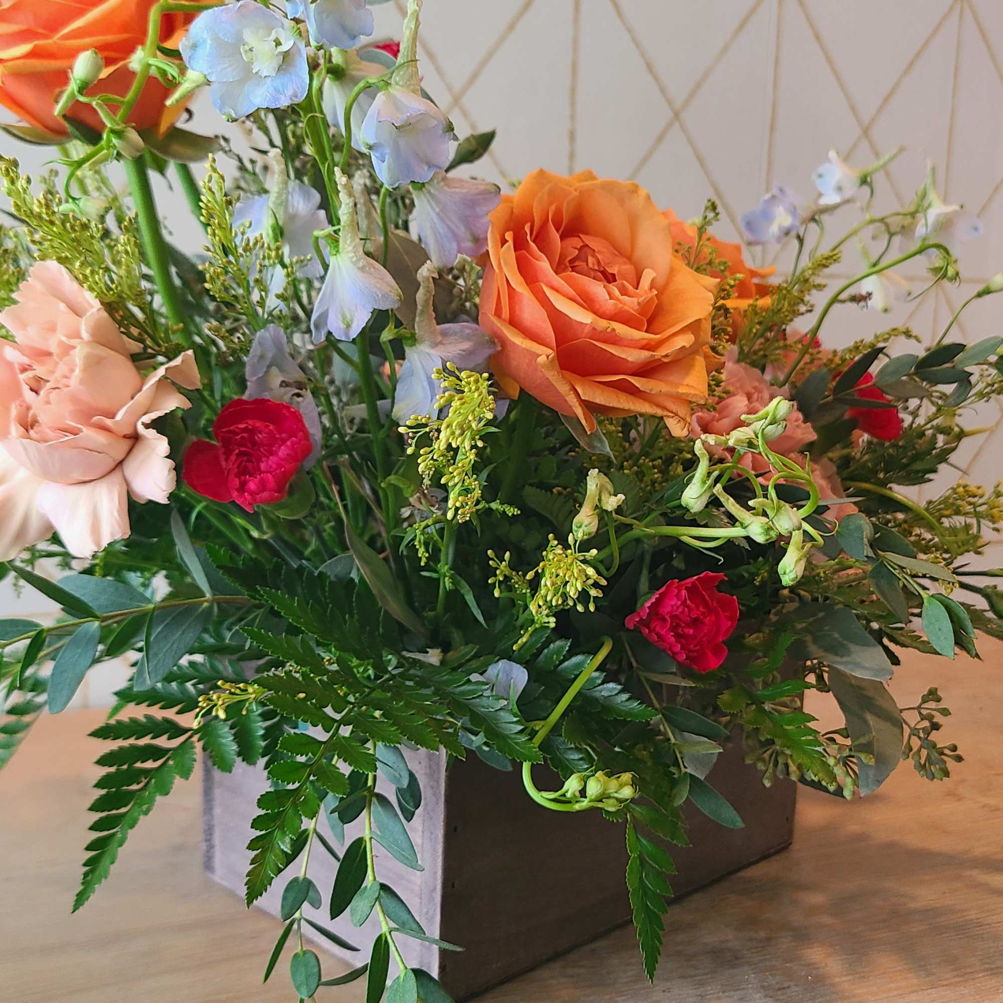 Mixed bouquet with orange roses, pale blue flowers, and pink carnations in a square vase